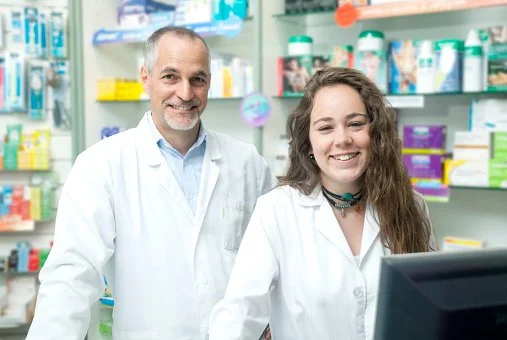 Two pharmacists in white coats standing in a pharmacy behind a counter, smiling at the camera.