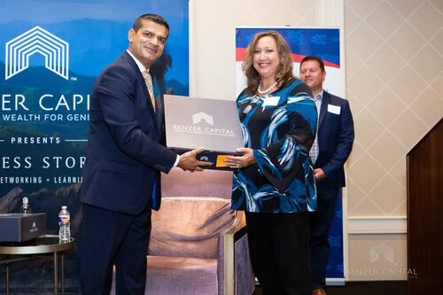 A man in a navy suit hands a box labeled 'Benzar Capital' to a woman in a blue patterned blouse, smiling, during a business event with banners and a third man in the background.