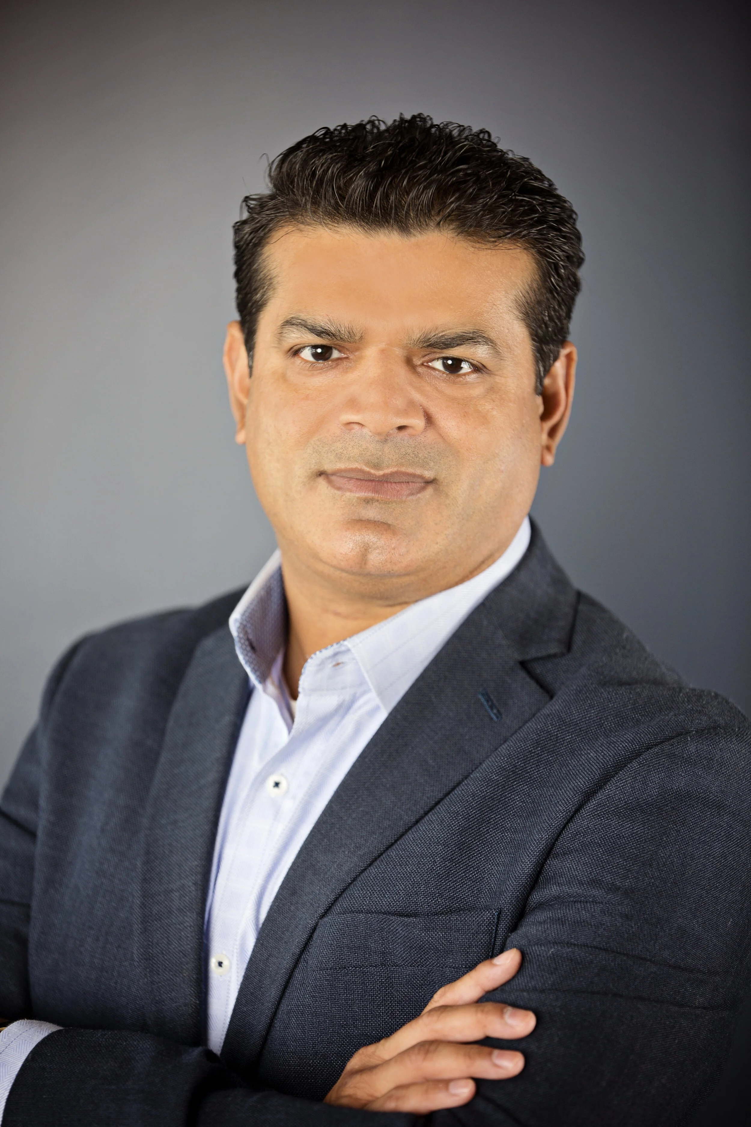 Professional portrait of a man in a dark suit jacket and light shirt, posing with arms crossed against a gray background.