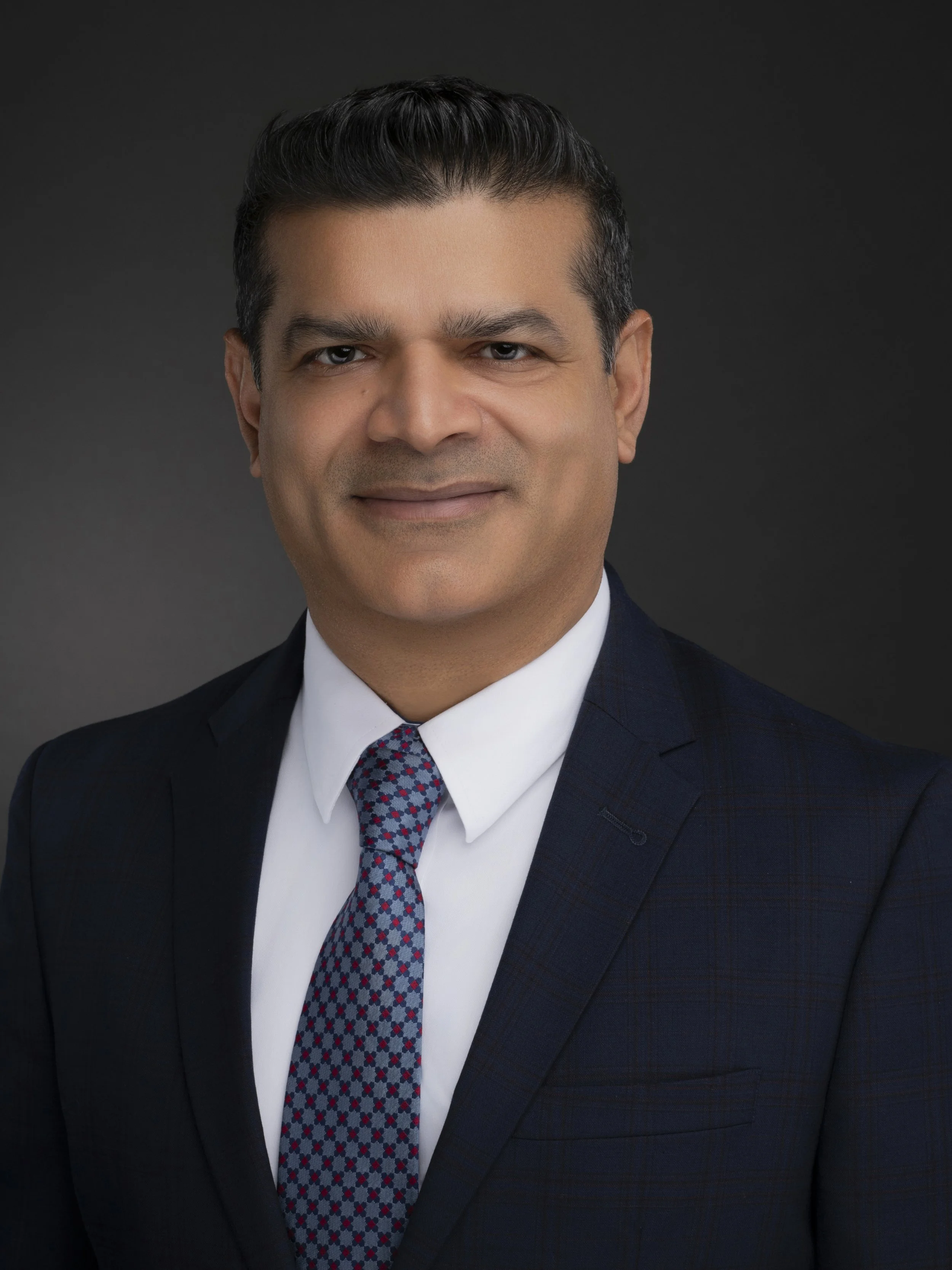 Professional portrait of a man in a dark suit jacket and light shirt, posing with arms crossed against a gray background.