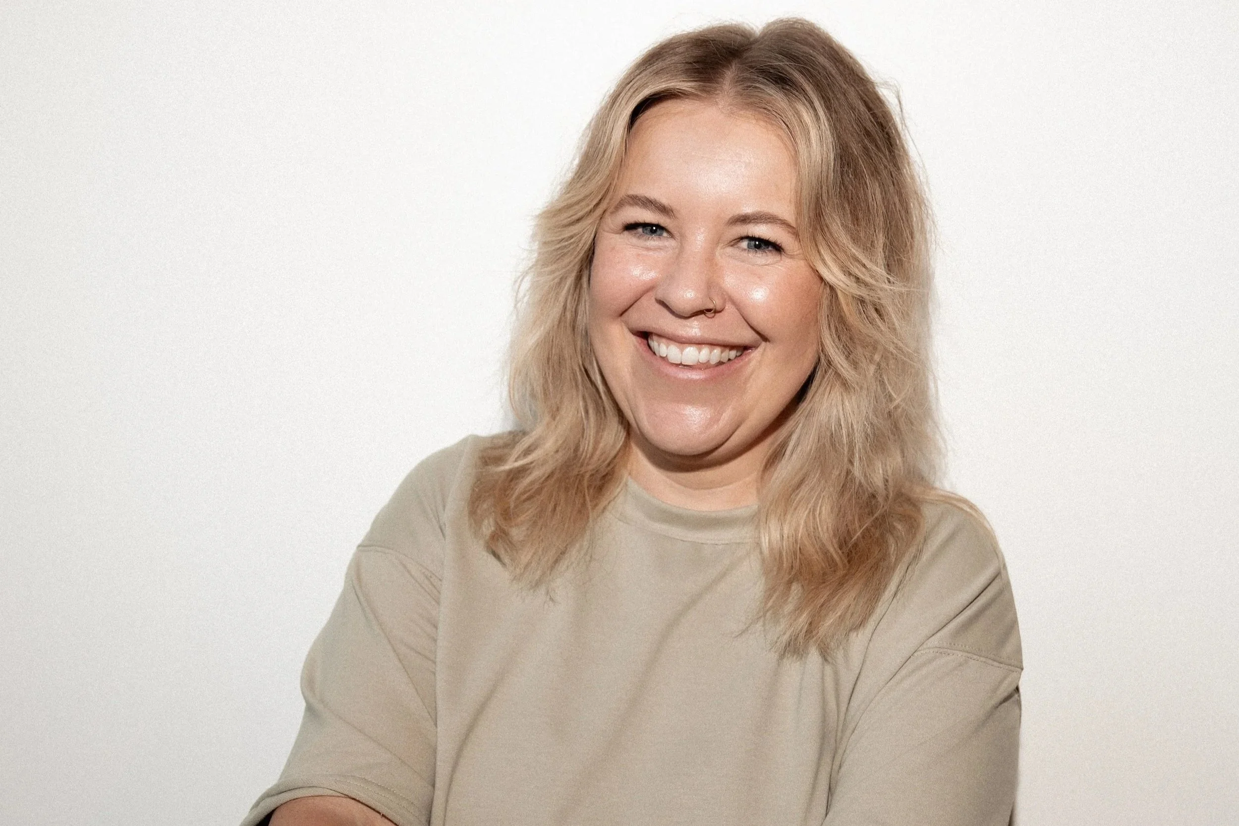 A smiling woman with short blonde hair, wearing a black blazer over a white top, standing against a plain beige wall.