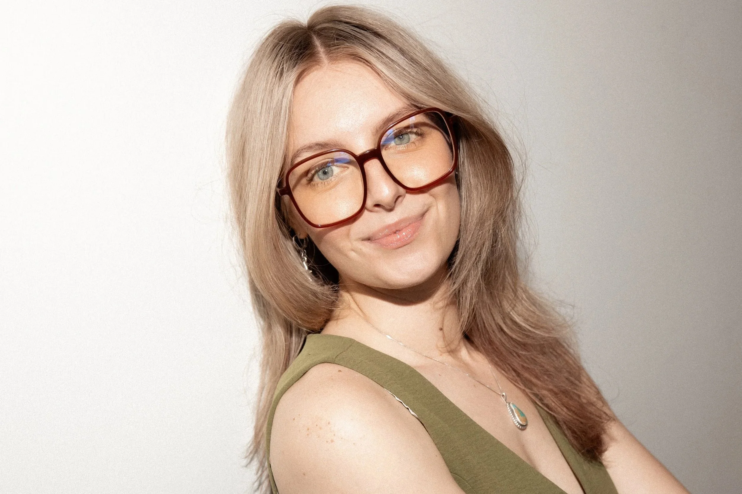 Young woman with long blonde hair and blue eyes, wearing a black sleeveless top, sitting against a white wall, with a small sun tattoo on her arm.