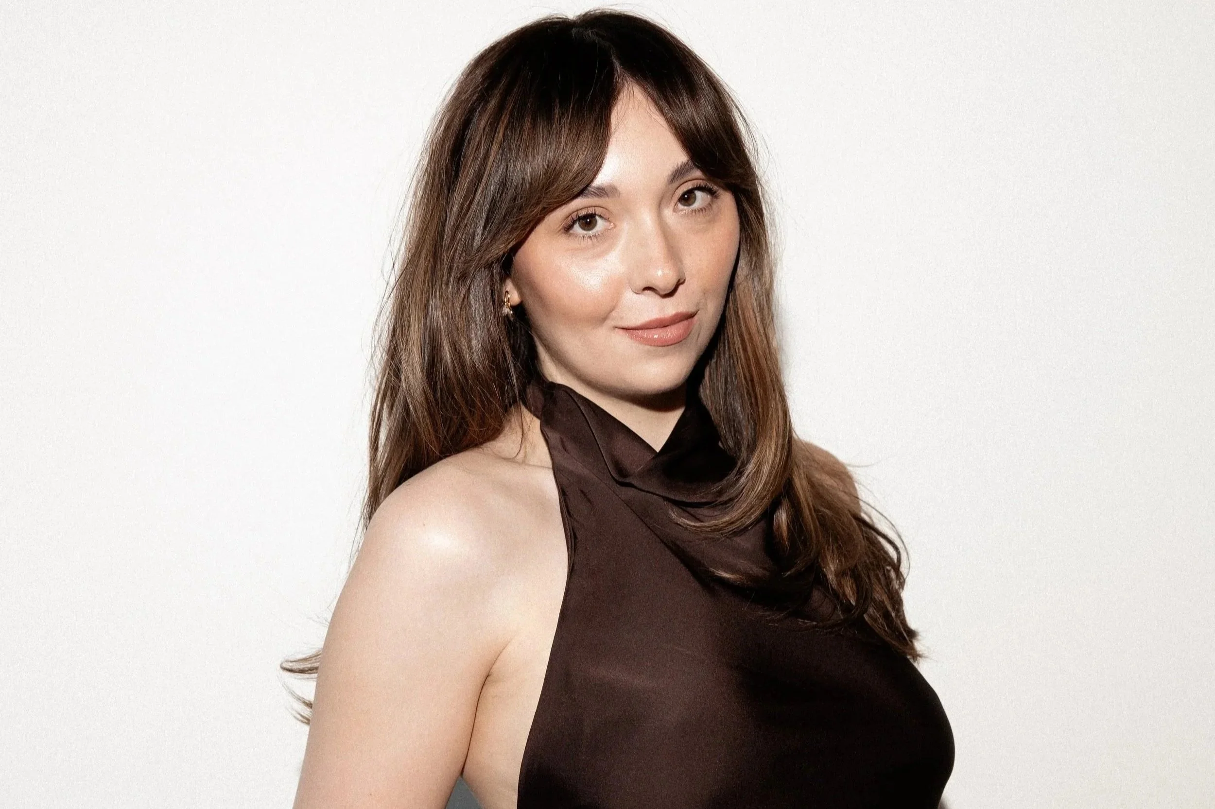 A woman with long, wavy brown hair wearing a black top, sitting against a plain white background.