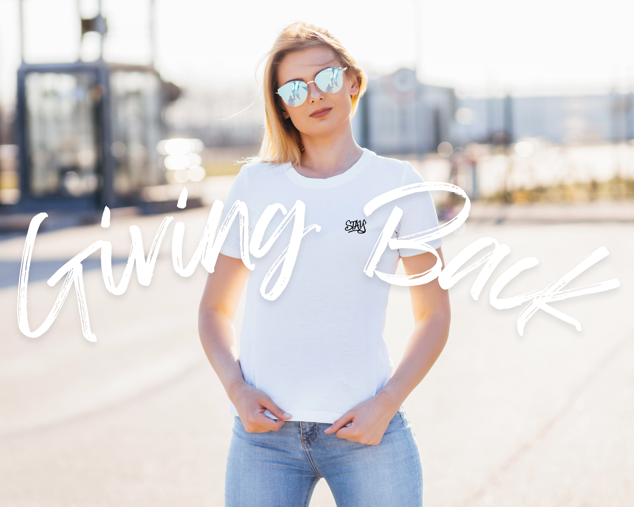 A young woman in sunglasses and a white t-shirt with the word stay standing outdoors with a blurred background of buses and buildings, with the words "Giving Back" in large white script overlaying the image.