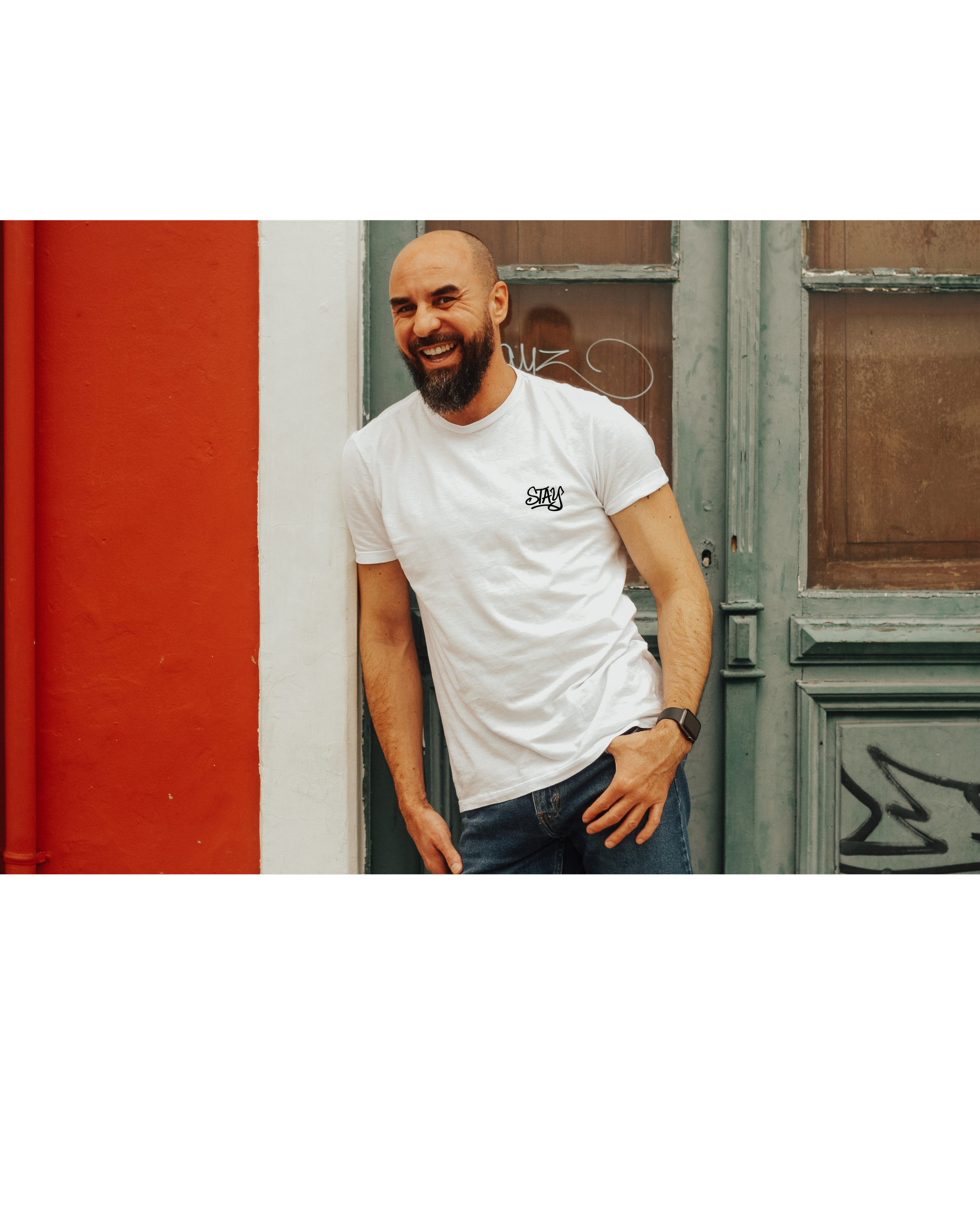 Young man with a beard smiling, wearing a white t-shirt with the word stay and jeans, standing outdoors against a colorful wall and old door. mental health