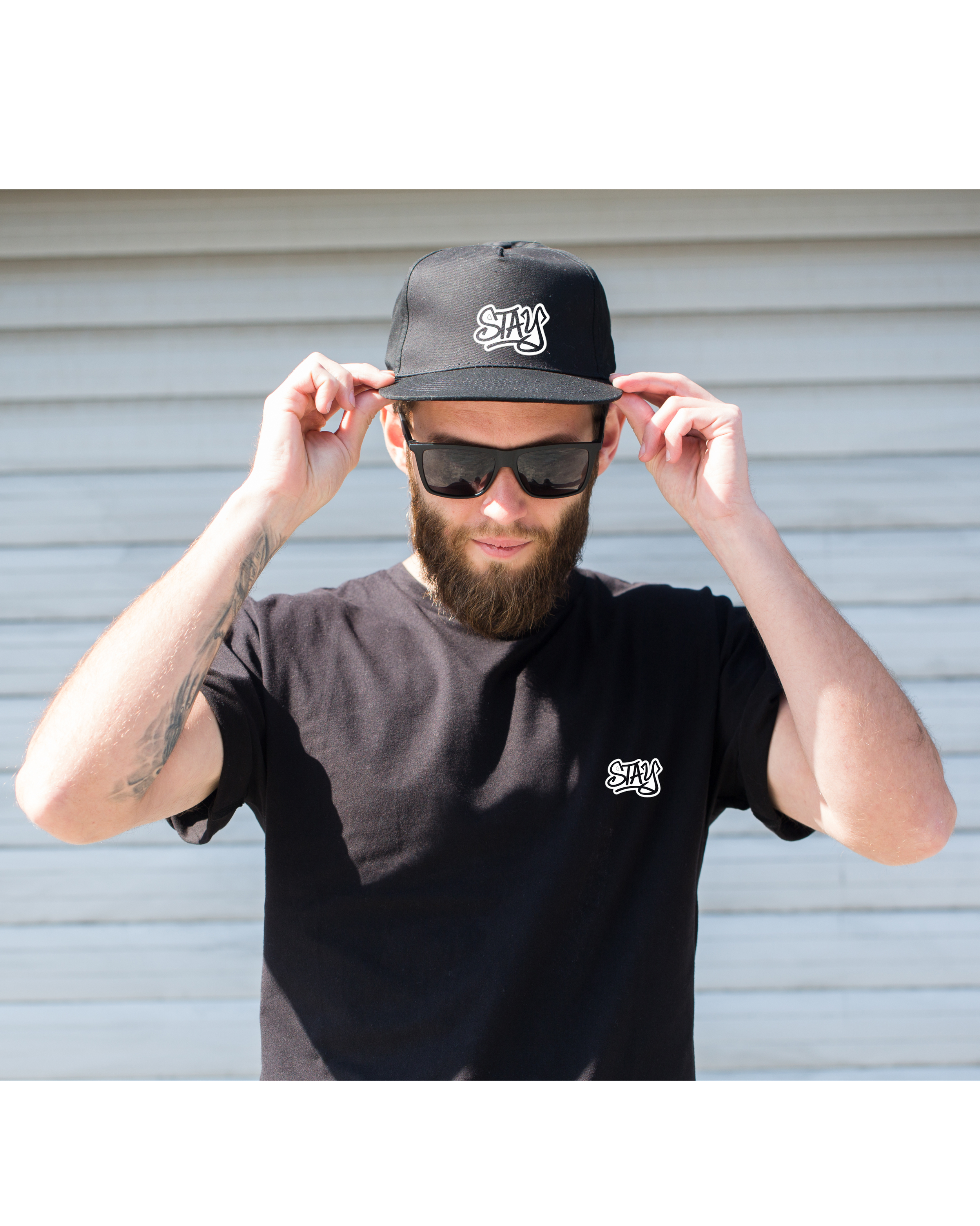 A man with a beard wearing black sunglasses and a black T-shirt, adjusting a black cap with the word 'STAY' embroidered on the front. Background features a horizontal metal siding.