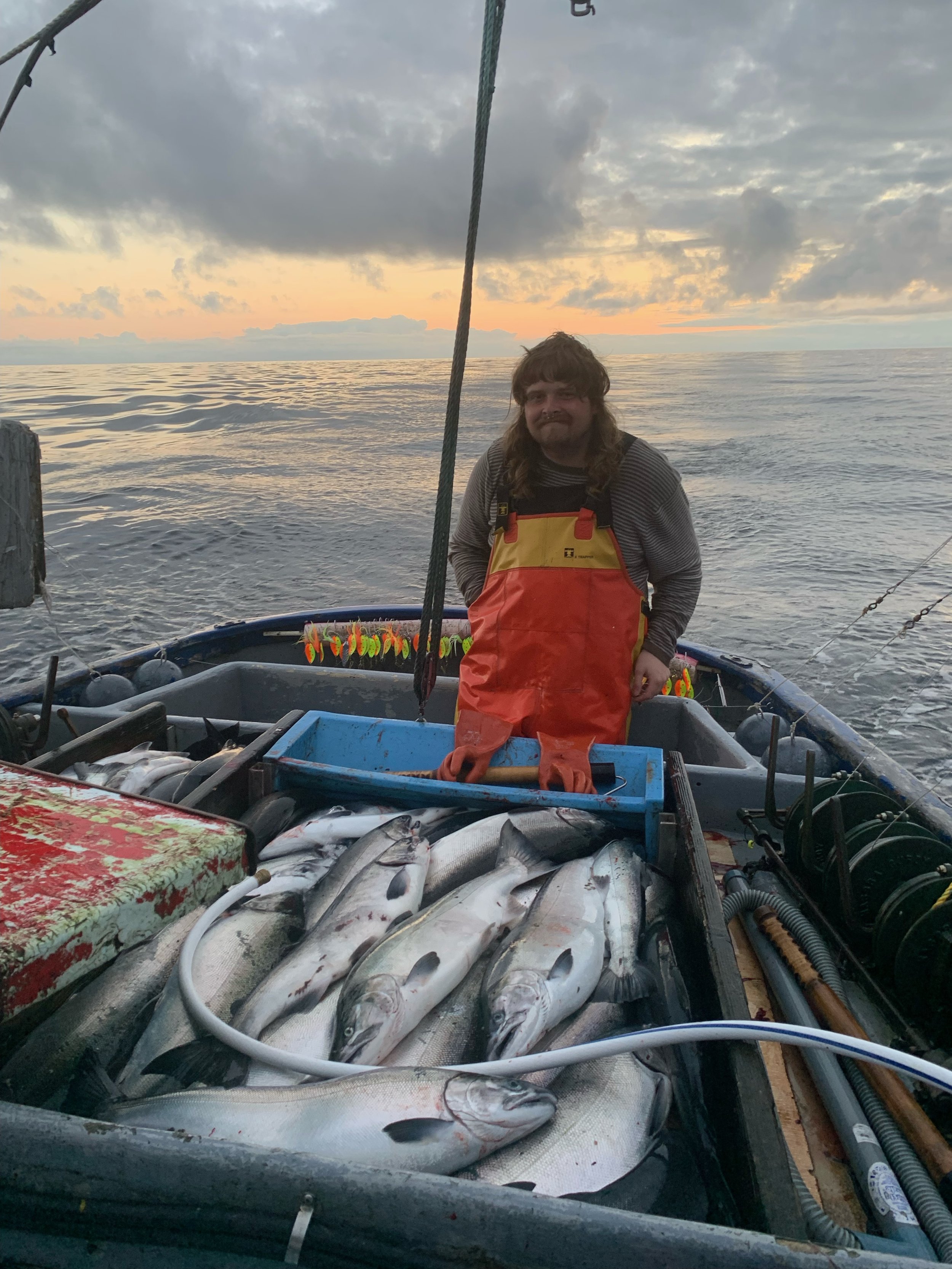 A person on a boat with caught fish, wearing orange waders, during sunset at sea.