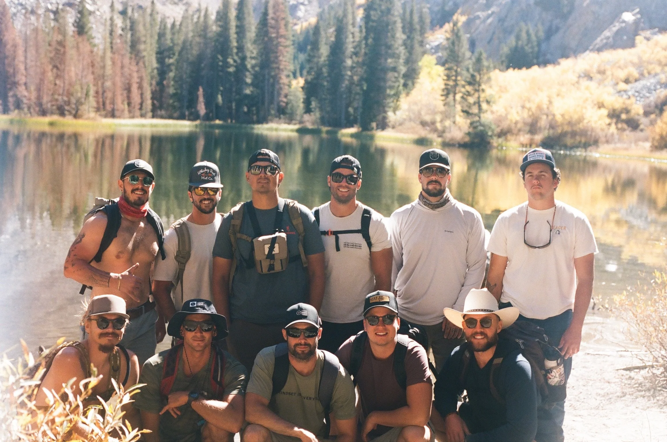 11 men standing by a lake surrounded by trees after a hike to the lake