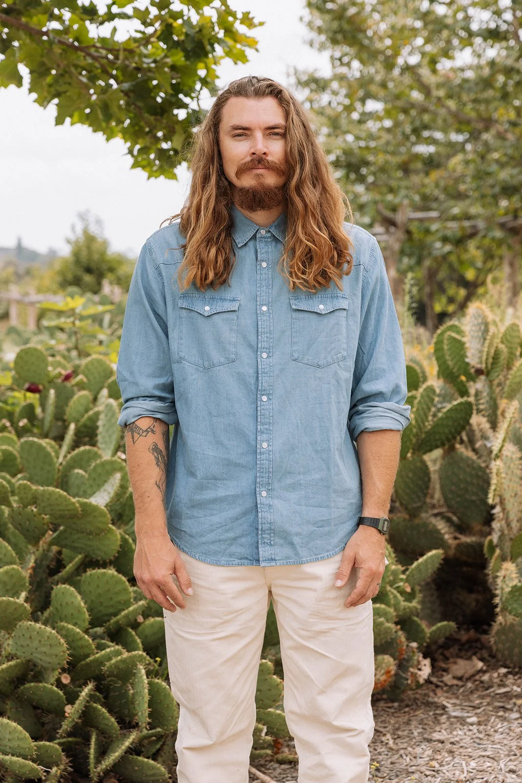 brock eissman standing in a blue button down shirt and white pants in a field surrounded by cactus. smiling with mouth closed looking at camera