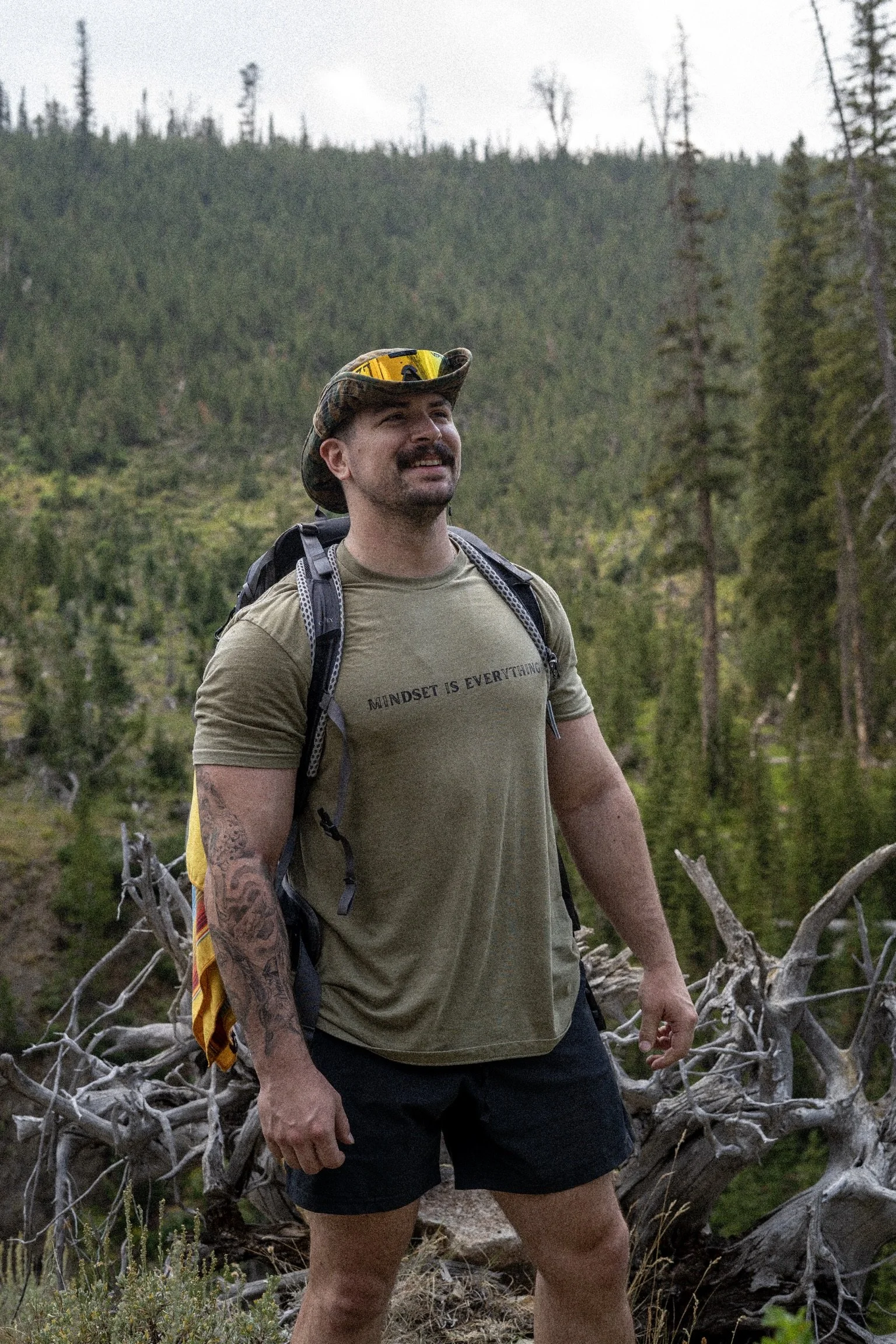 MASON STANDING IN A GREEN MINDSET IS EVERYTHING SHIRT WEARING A CAMO HAT WITH SUNGLASSES SURROUNDED BY TREES