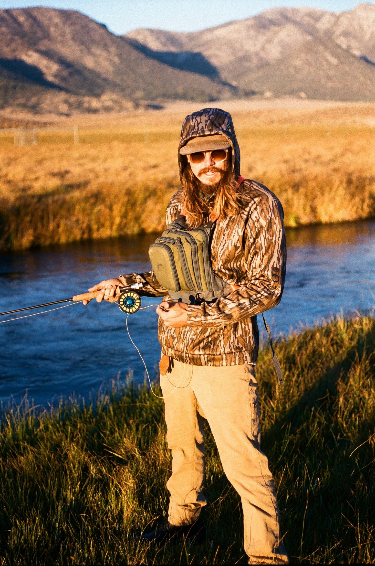 brock in camo hoodie with a fly rod standing next to the river fishing