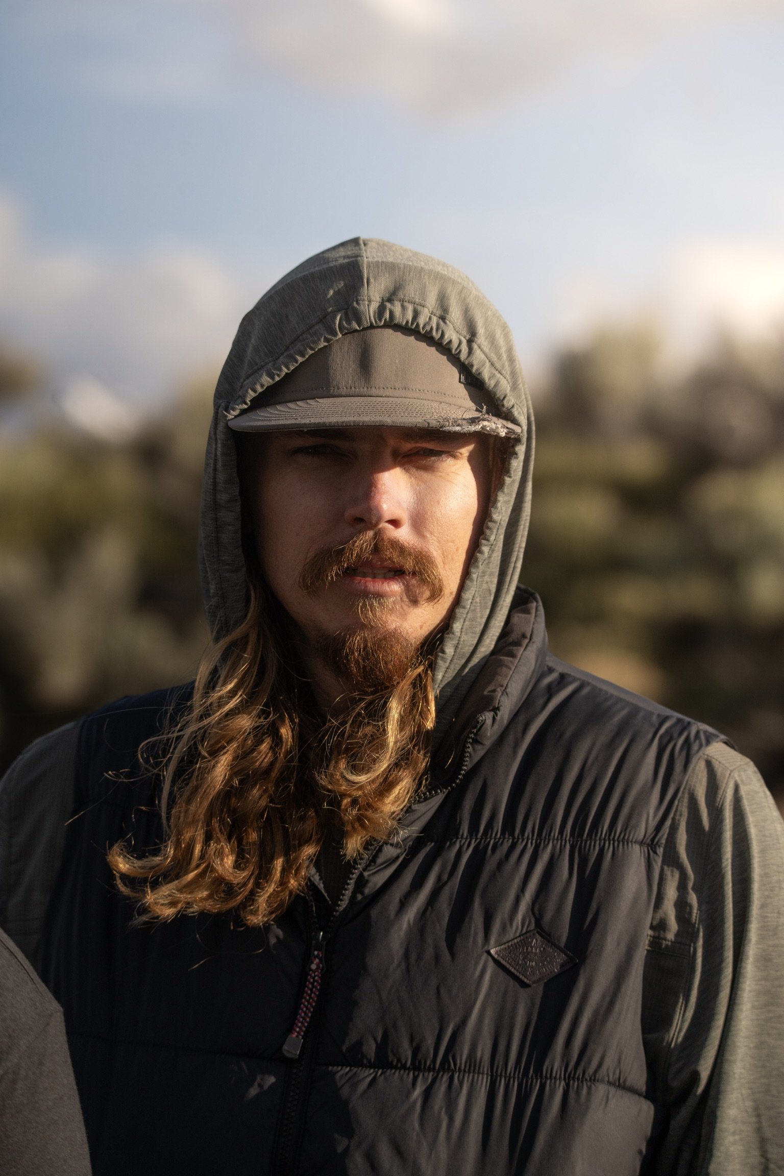 Brock closeup shot of him wearing a green and grey hoodie with a hat glaring at the camera