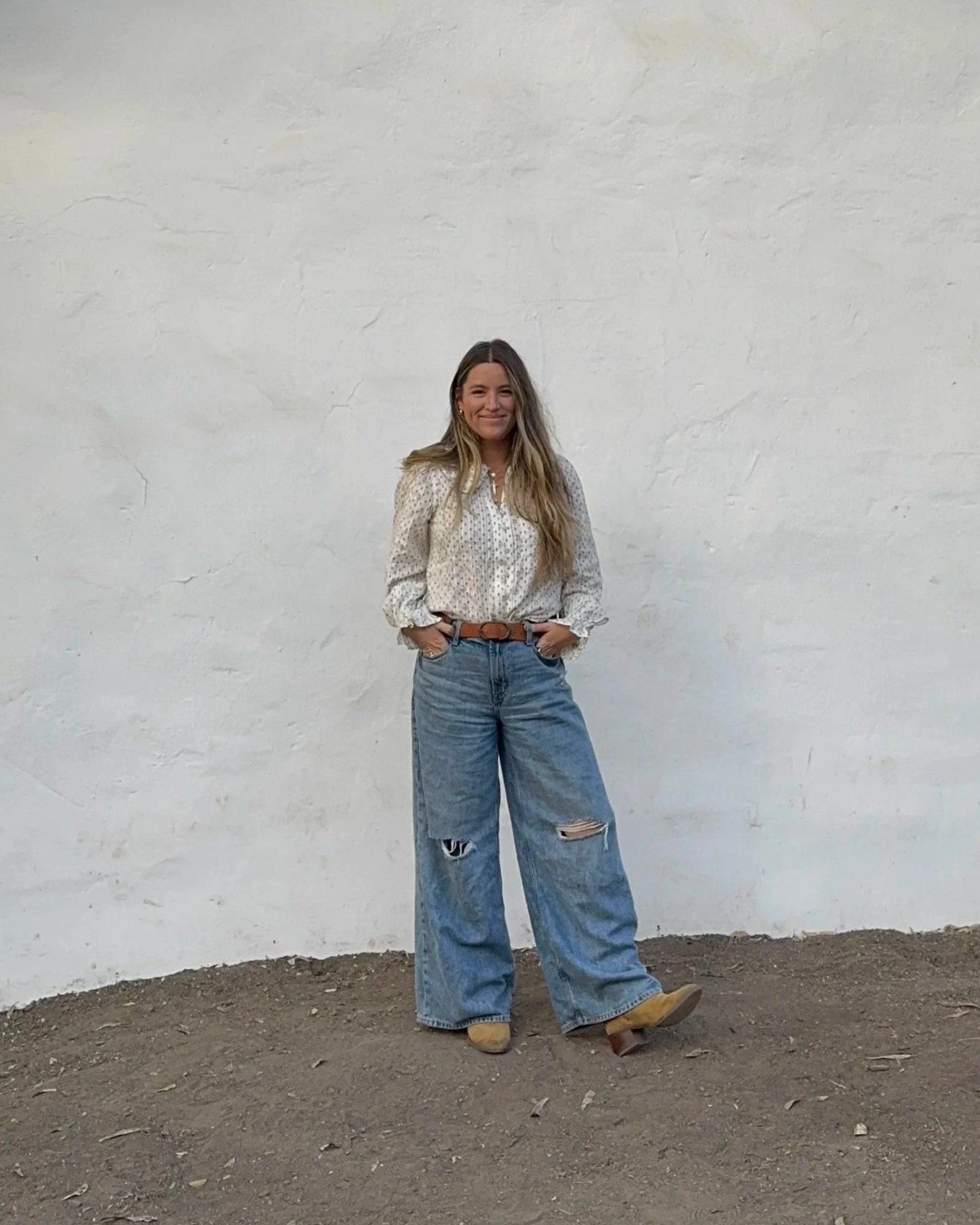 Bree Eissman standing against white wall wearing blue jeans, floral blouse and smiling.