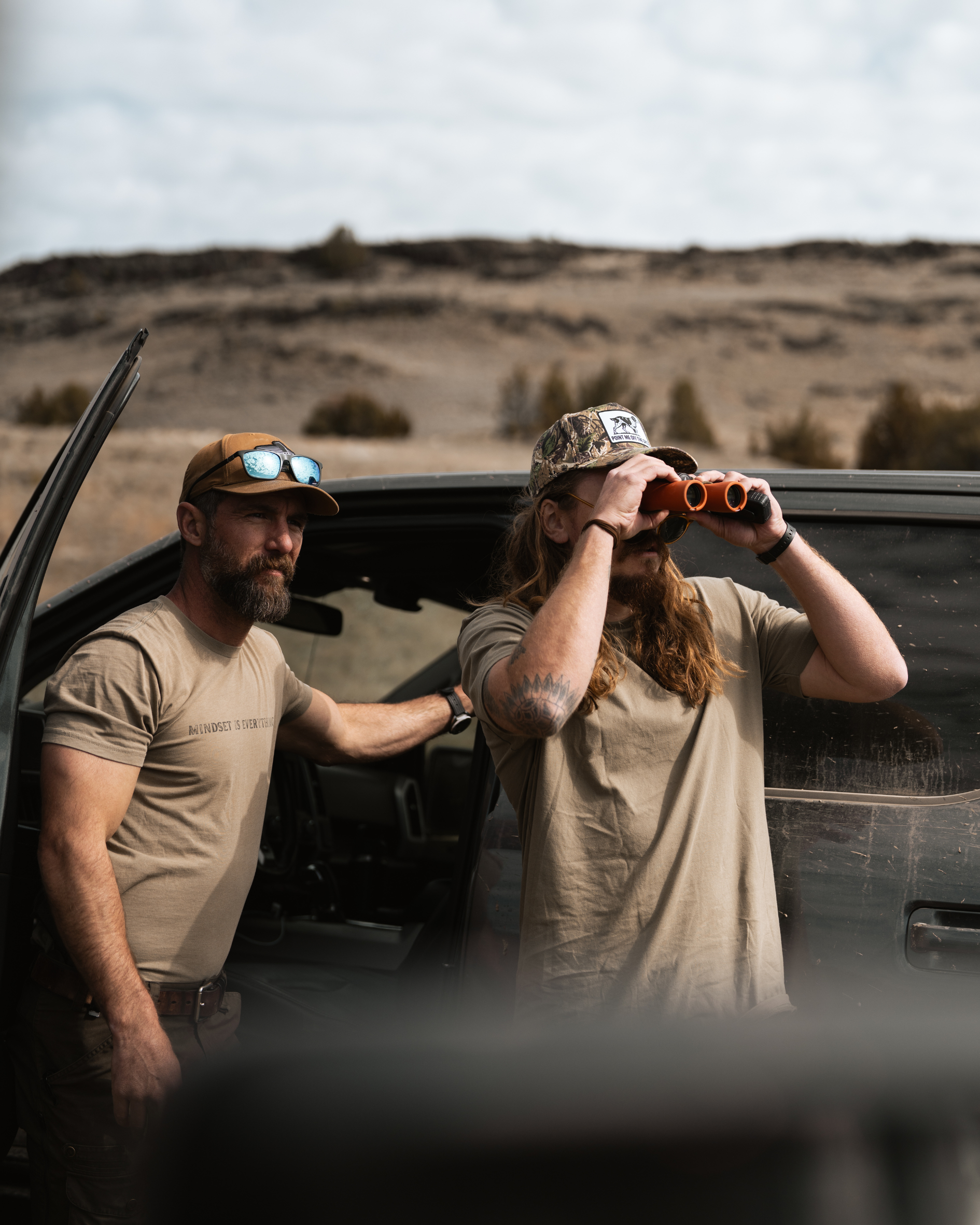 2 men standing near truck using binoculars looking in to the far distance. both are wearing green off the gryd shirts with baseball hats