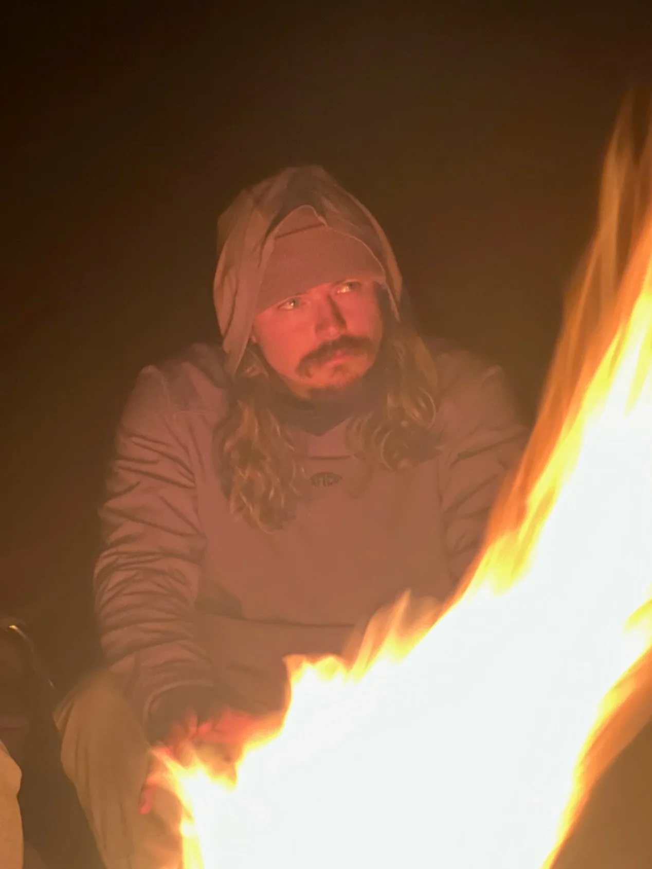 MAN LOOKING INTENSELY NEXT TO CAMPFIRE WEARING A HOODIE