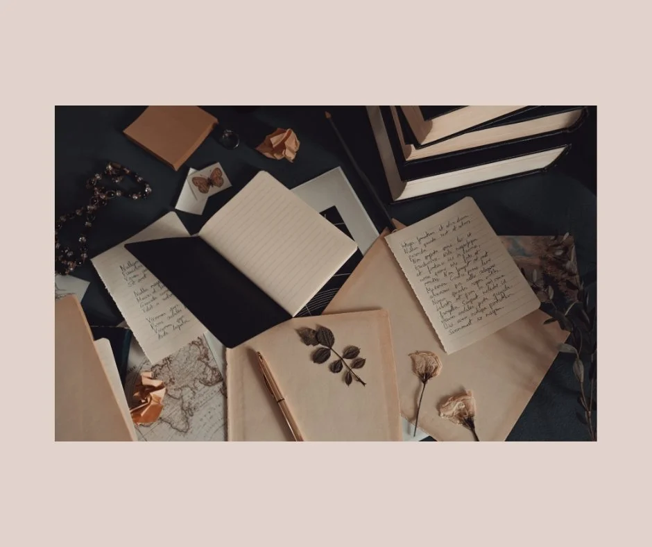 Open notebooks, handwritten notes, dried flowers, a pen, a map, jewelry, and stacked black binders on a dark surface.