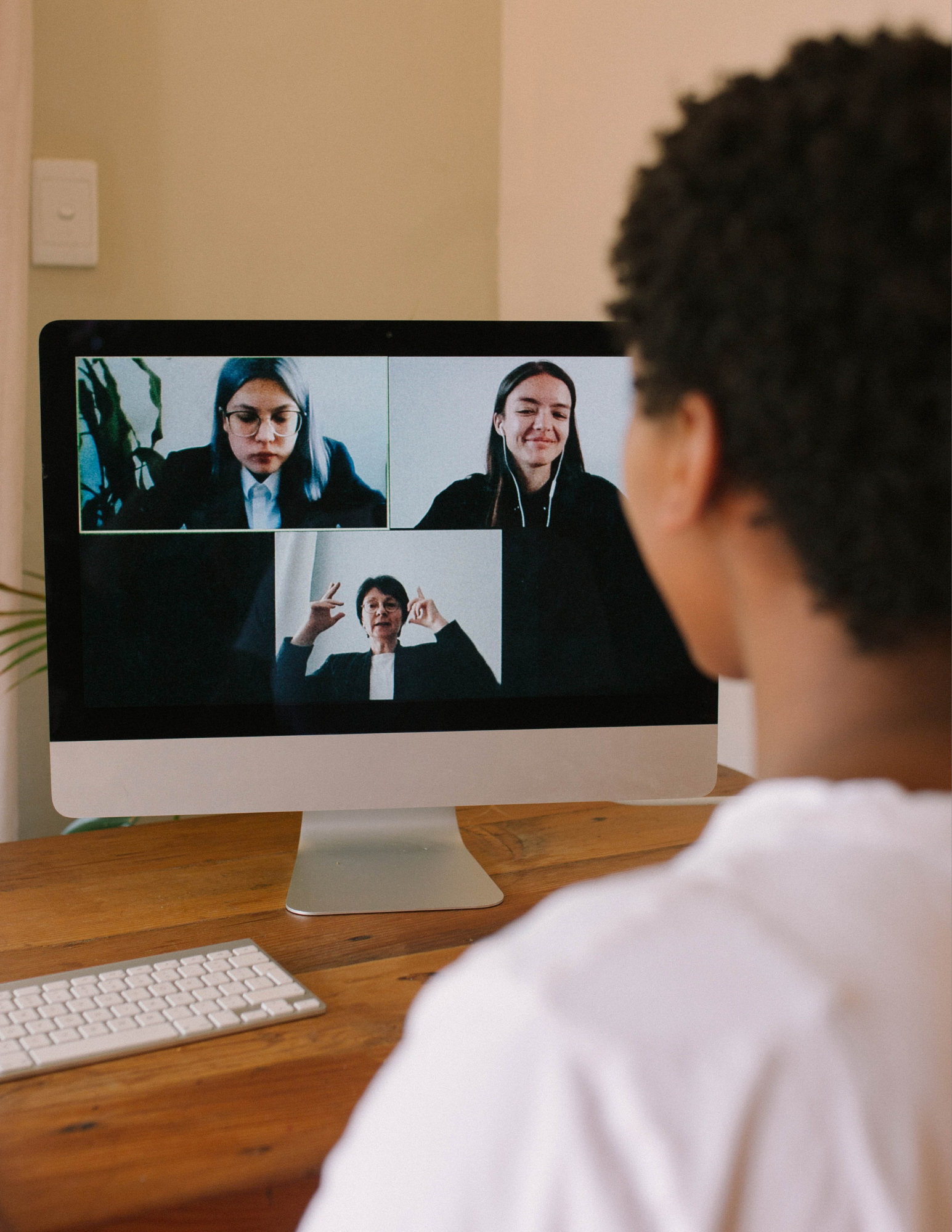 Person participating in a virtual video conference call on a desktop computer, with three women visible on the screen in a grid layout. The person is sitting at a wooden desk, viewed from behind, wearing a white shirt.