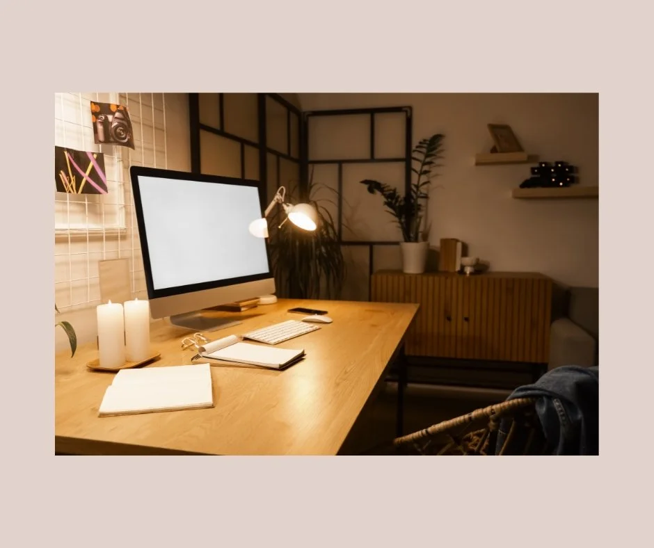 A cozy home office with a wooden desk, desktop computer, candles, notebooks, and a desk lamp, with plants and shelves in the background.