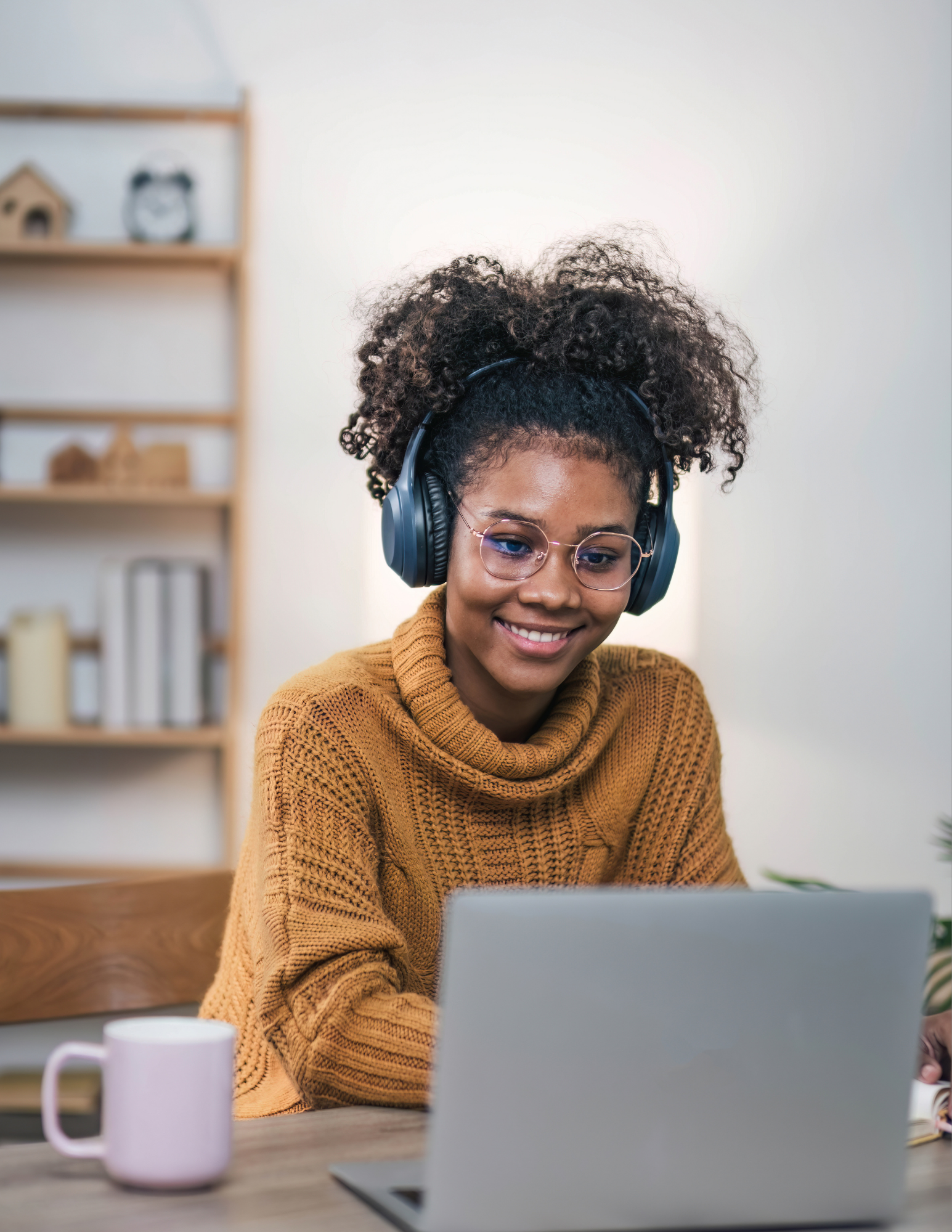 A woman with curly hair, wearing glasses and a mustard sweater, sitting at a desk with a laptop, listening to headphones and smiling.