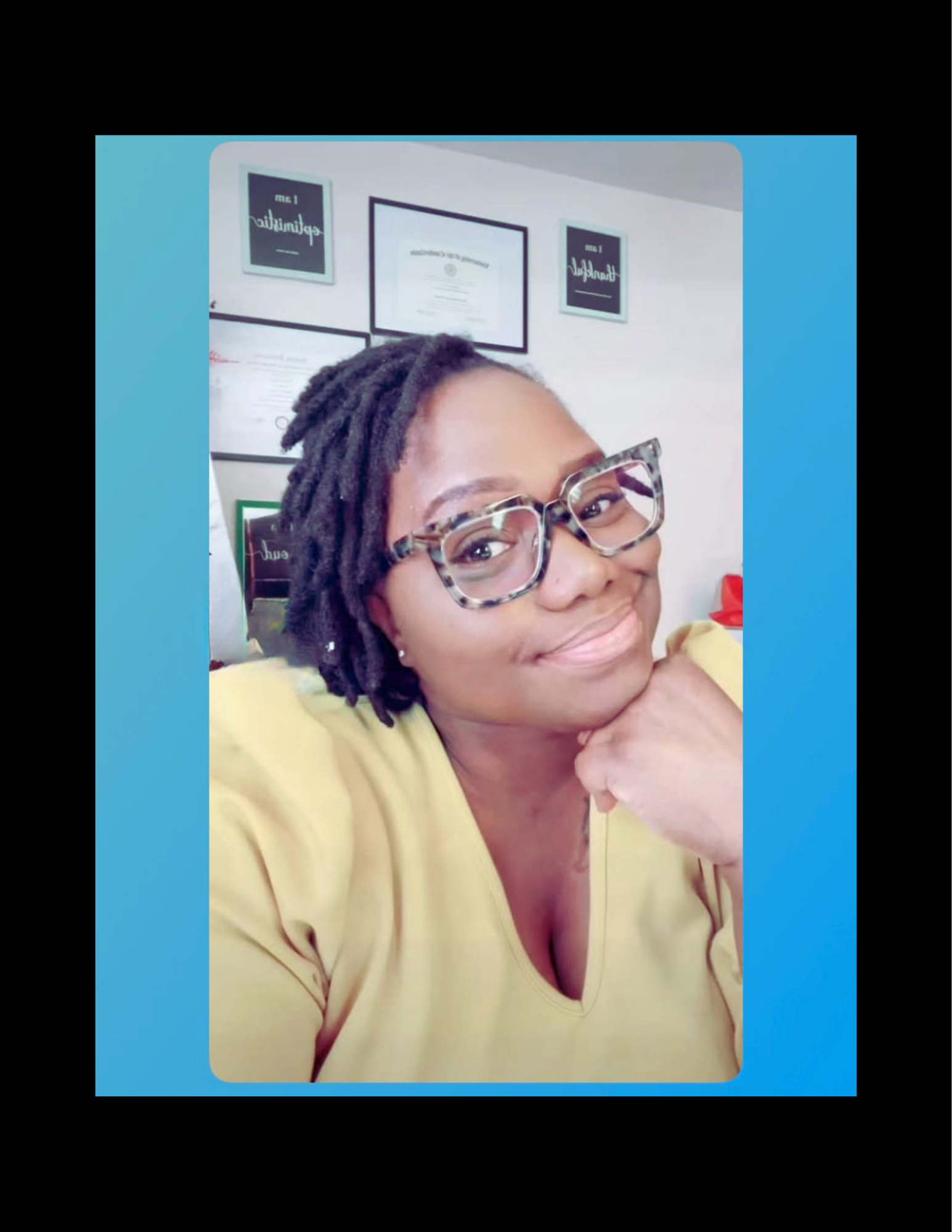 A woman with short dreadlocks, wearing glasses and a yellow top, smiling and resting her chin on her hand in an office setting.