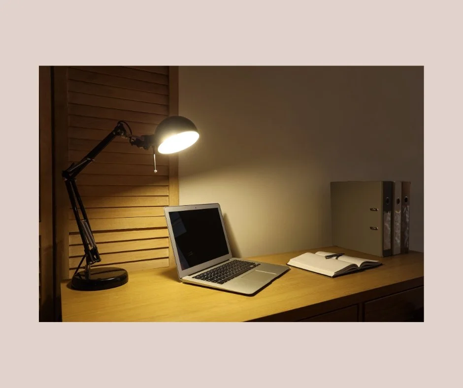A wooden desk with a black desk lamp, a silver laptop, a notebook, a pen, and three storage folders, with a louvered wooden partition behind.