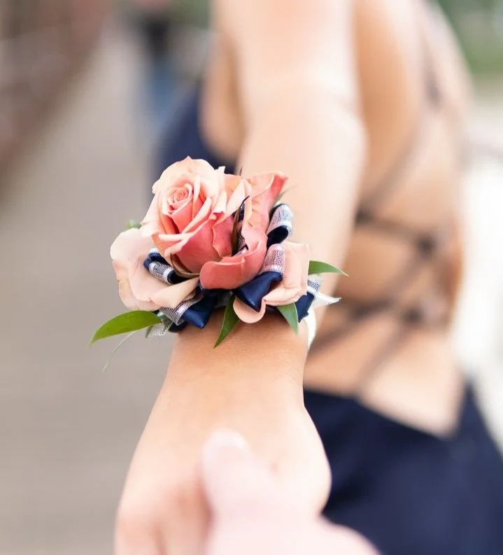 A person wearing a wrist corsage with pink roses, blue ribbons, and green leaves, holding another hand.