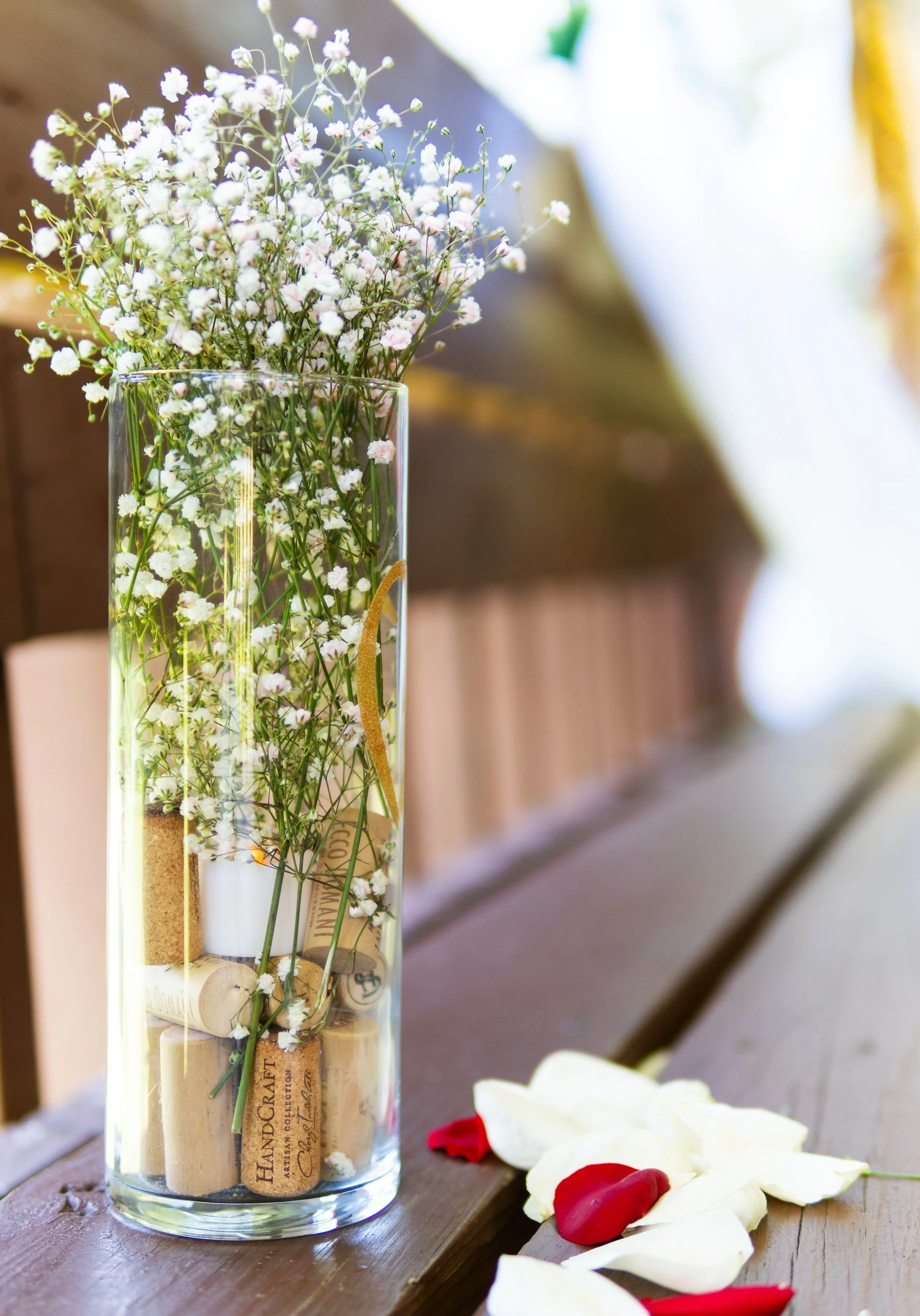 A tall glass vase filled with white baby's breath flowers and wine corks, placed on a wooden table with white and red flower petals scattered nearby.