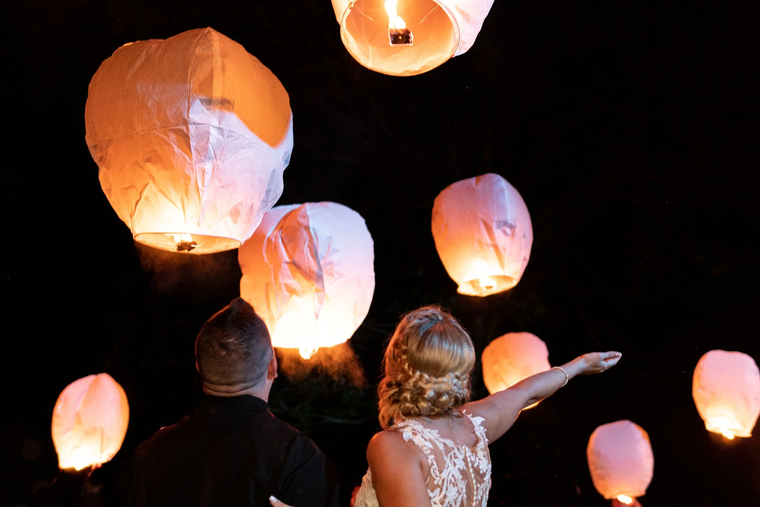 A couple, possibly newlyweds, releasing sky lanterns at night. The man has short dark hair and is wearing a dark suit, while the woman has blonde hair styled in curls and is wearing a white lace dress. The sky is dark, and several lit sky lanterns are floating upward around them.