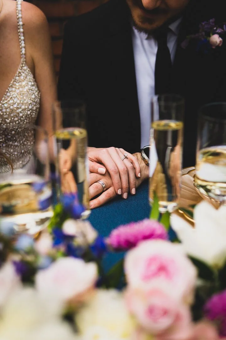 Close-up of a couple holding hands with wedding rings, seated at a table with glasses of champagne, surrounded by colorful flowers.