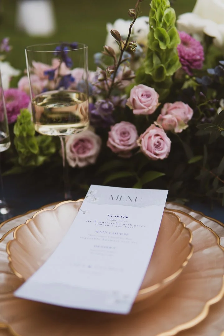Elegant table setting featuring a menu card on a scalloped gold plate, surrounded by pink roses, purple flowers, a glass of white wine, and a floral centerpiece with various blooms.