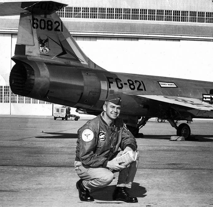 Black and white photo of a smiling pilot in uniform squatting in front of an F-104 jet aircraft on an airfield.