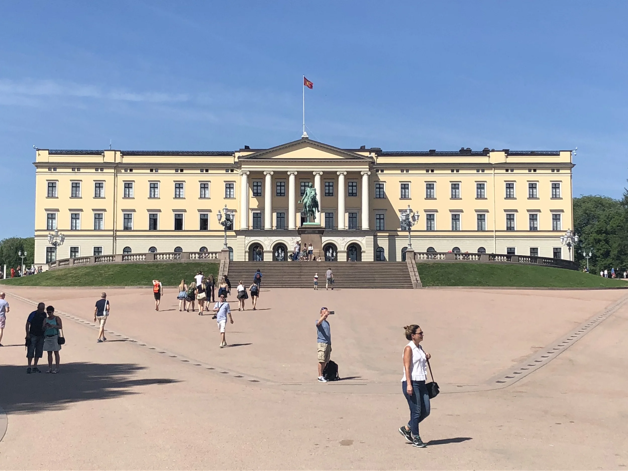 Large yellow building with a central archway, columns, and a flag on top. Front steps lead up to it with visitors walking around, and a statue of a person on horseback in front.