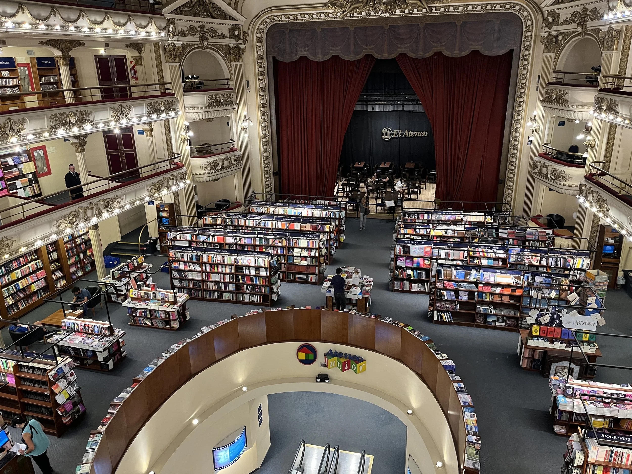Interior view of a grand bookstore or library with ornate architecture, multiple bookshelves filled with books, an elevated balcony, a stage with red curtains, and people browsing.