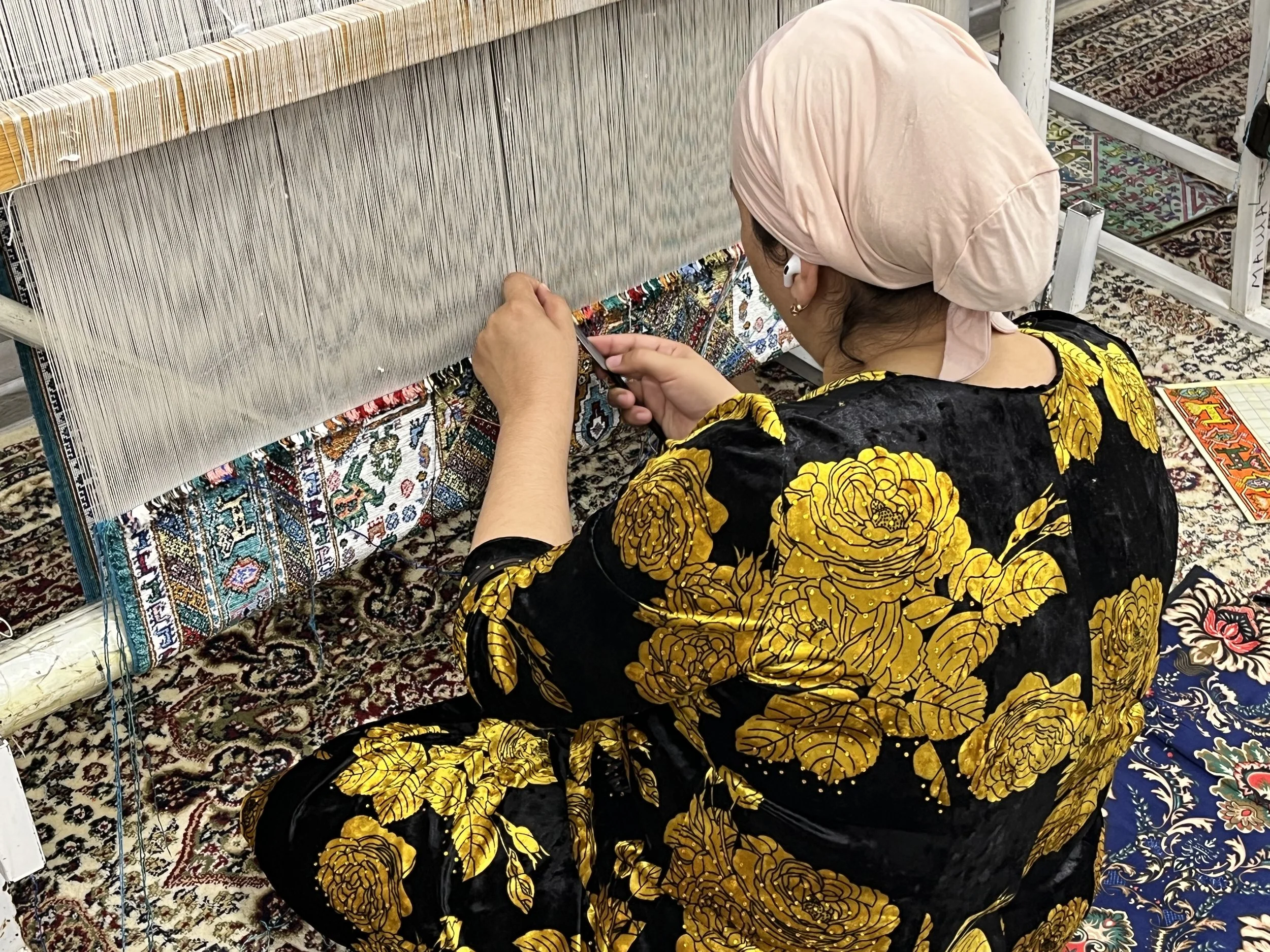 Woman weaving in a textile workshop, wearing a pink headscarf and a black and yellow floral dress, sitting on a patterned rug and working on a traditional loom.