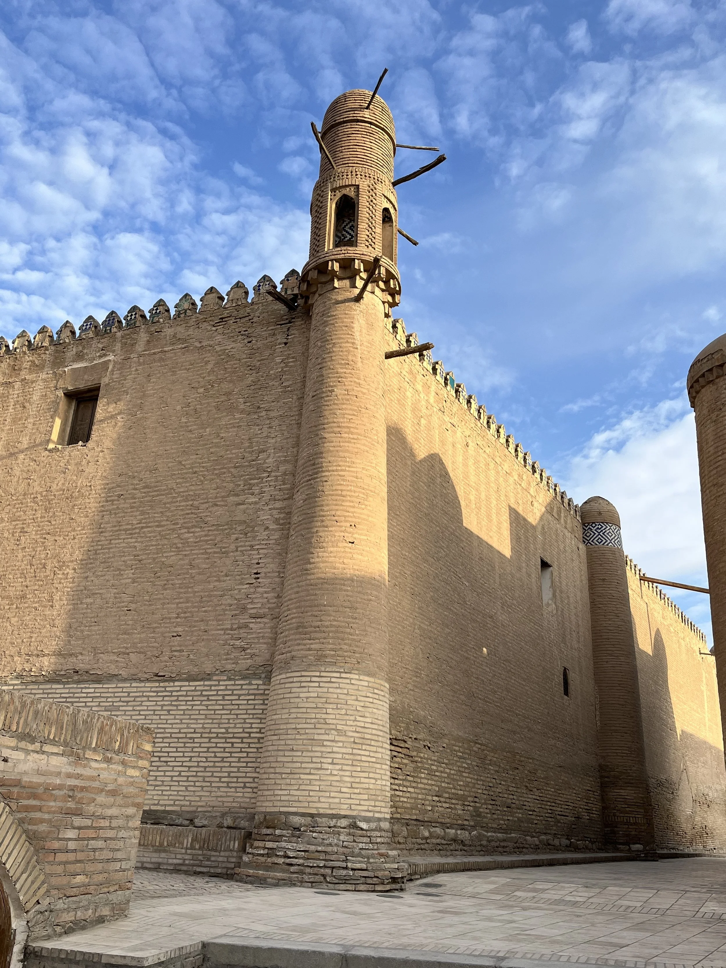 An old brick fortress with a tall, rounded tower and wooden sticks protruding from the top, against a blue sky with scattered clouds.