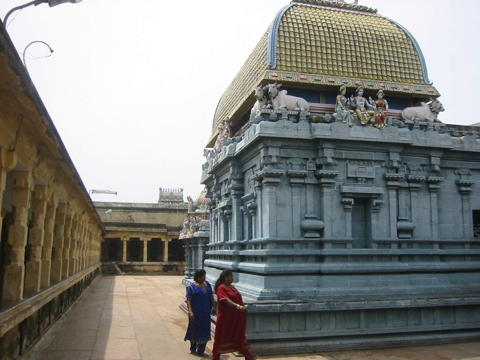 Two women in traditional Indian sarees walking past a large Hindu temple with intricate carvings and a golden domed roof.