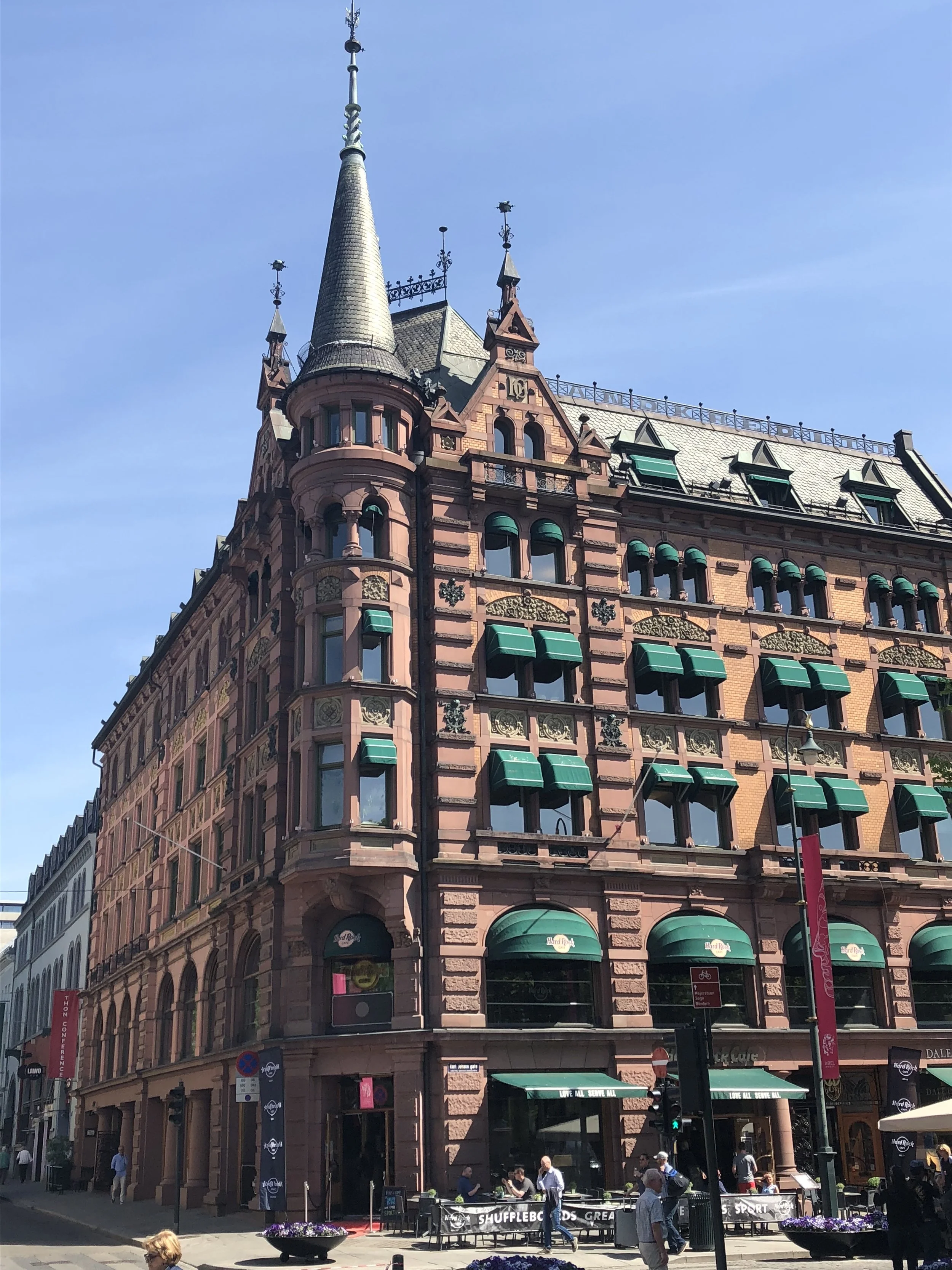 A historic pink stone building with green awnings and a turret, located on a busy city street with pedestrians, outdoor seating, and a clear blue sky.