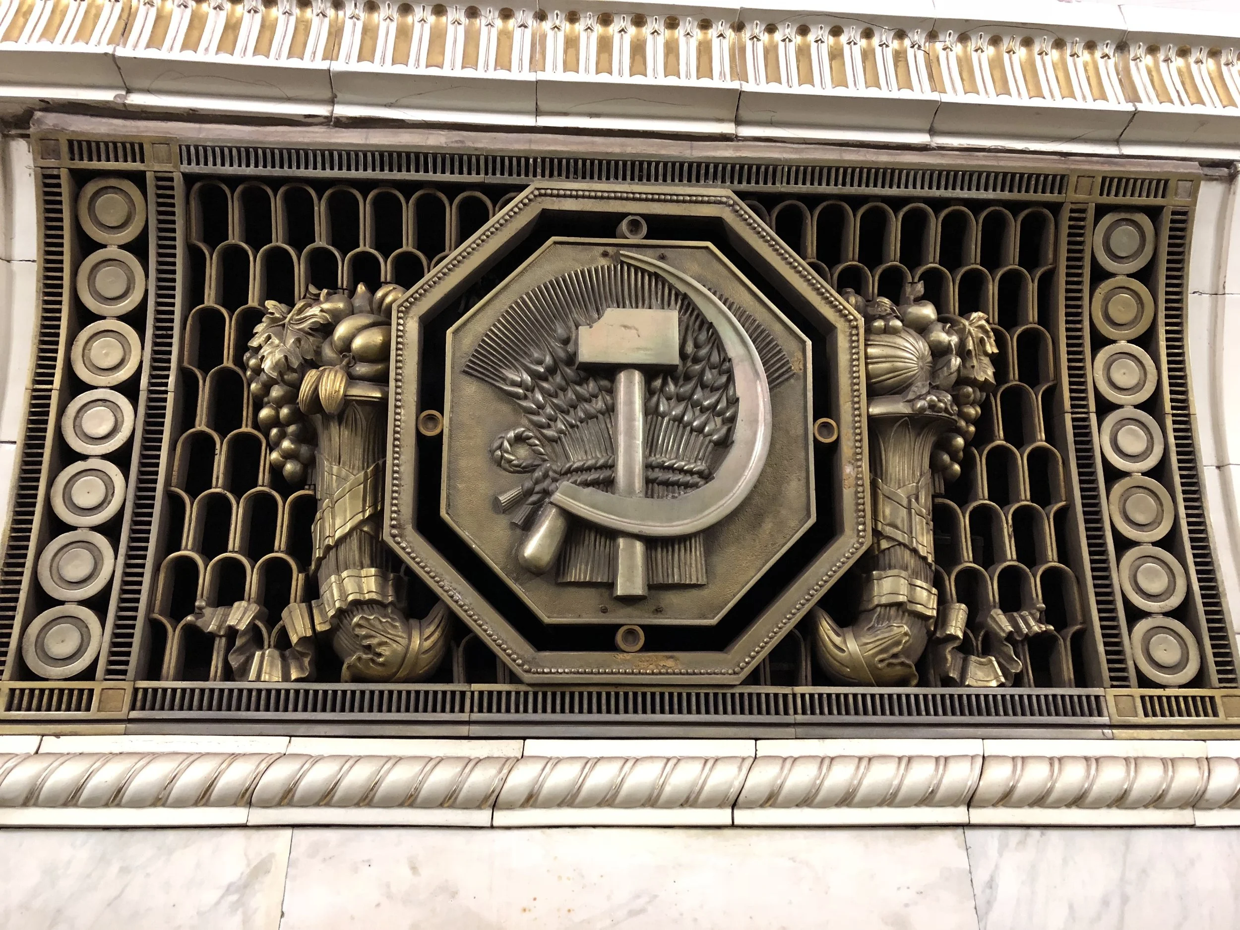 A bronze or brass emblem of the Soviet Union, featuring a hammer and sickle with wheat ears, mounted on a decorative grate with circular pattern and surrounded by ornate lion or dragon figures, located in an indoor setting.