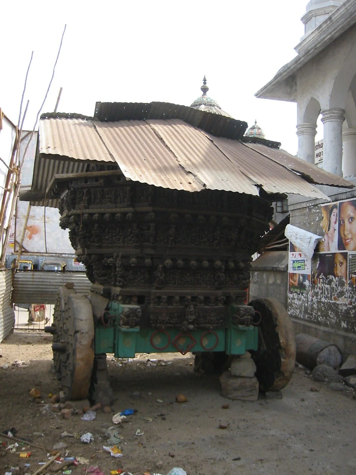 A rustic, wooden cart with a corrugated metal roof, possibly used for transportation or ceremonial purposes, situated outdoors near buildings and posters on a wall.