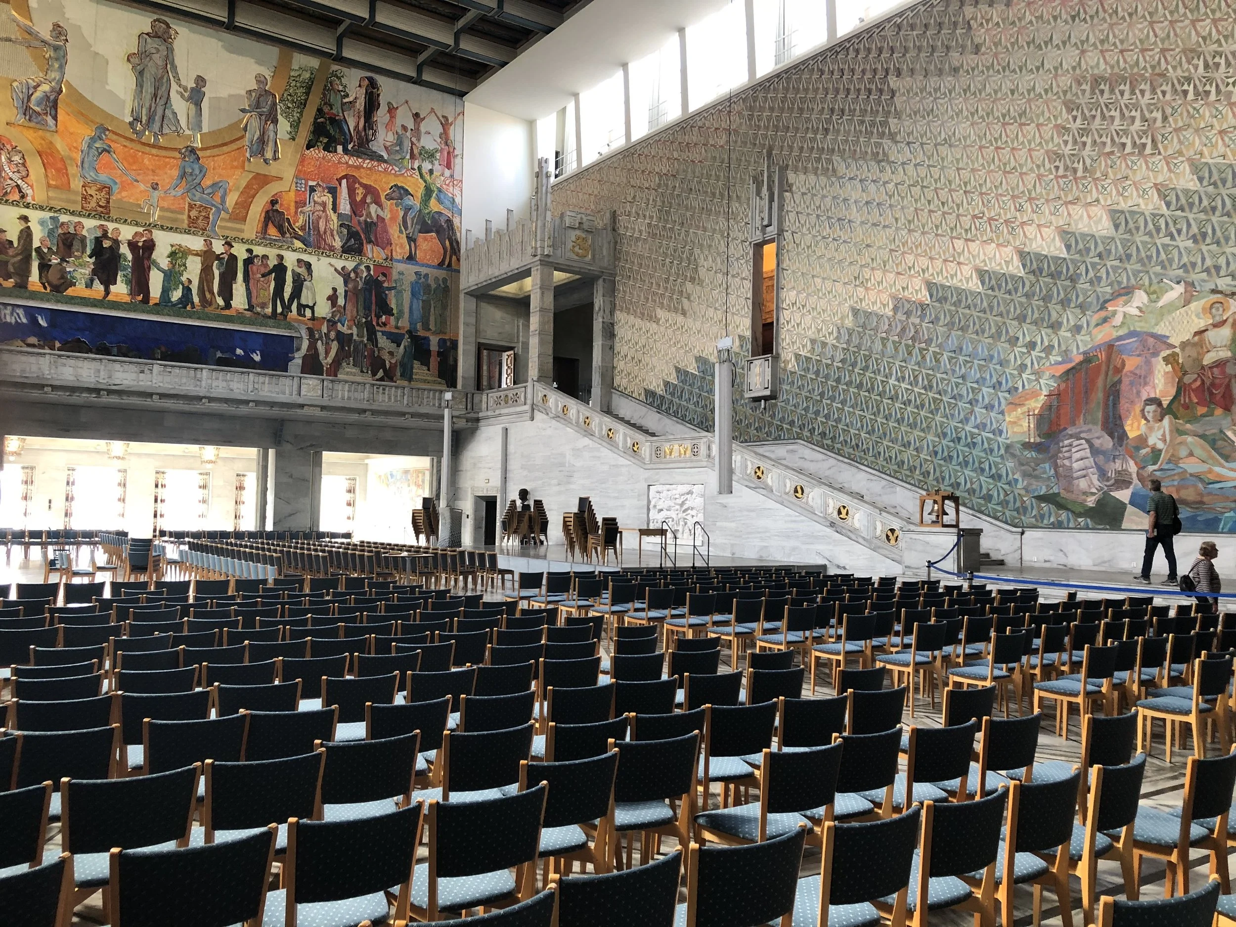 An auditorium with multiple rows of chairs facing a stage, with ornate murals on the walls depicting historical or cultural scenes, and a marble staircase leading to an upper level.