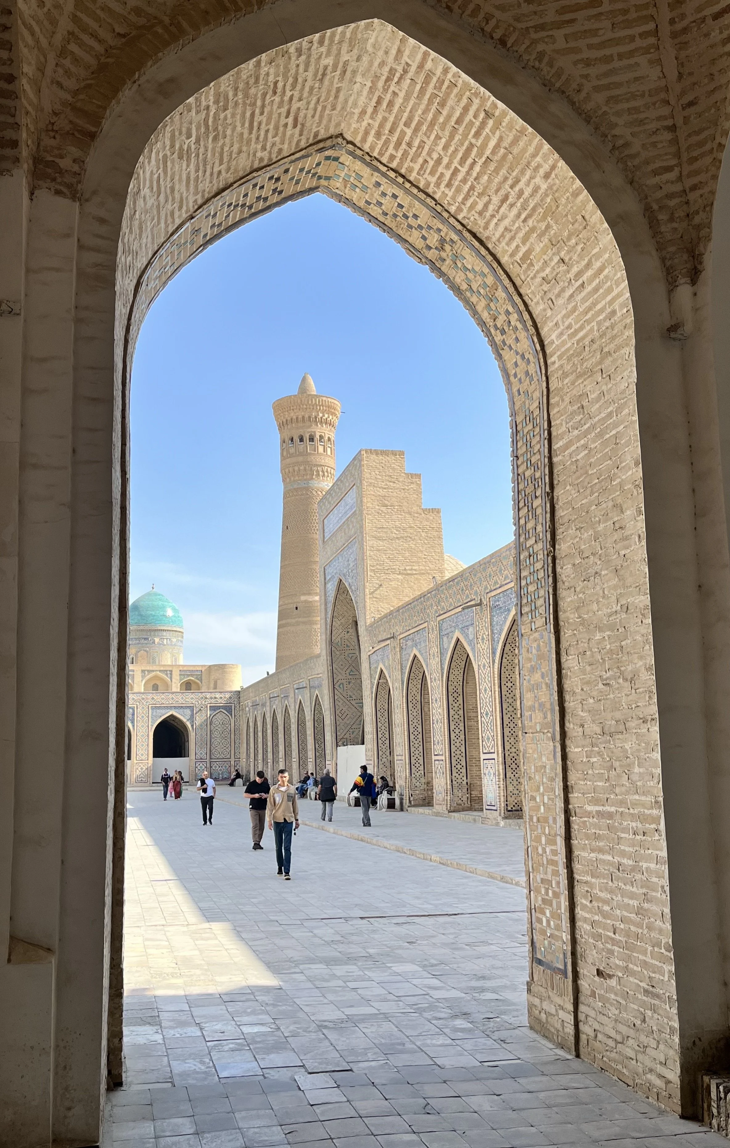 View of a historic Islamic architecture courtyard with arched doorways, a tall minaret, and a dome with a turquoise top, seen through a decorative brick archway, with visitors walking on the paved courtyard under a blue sky.