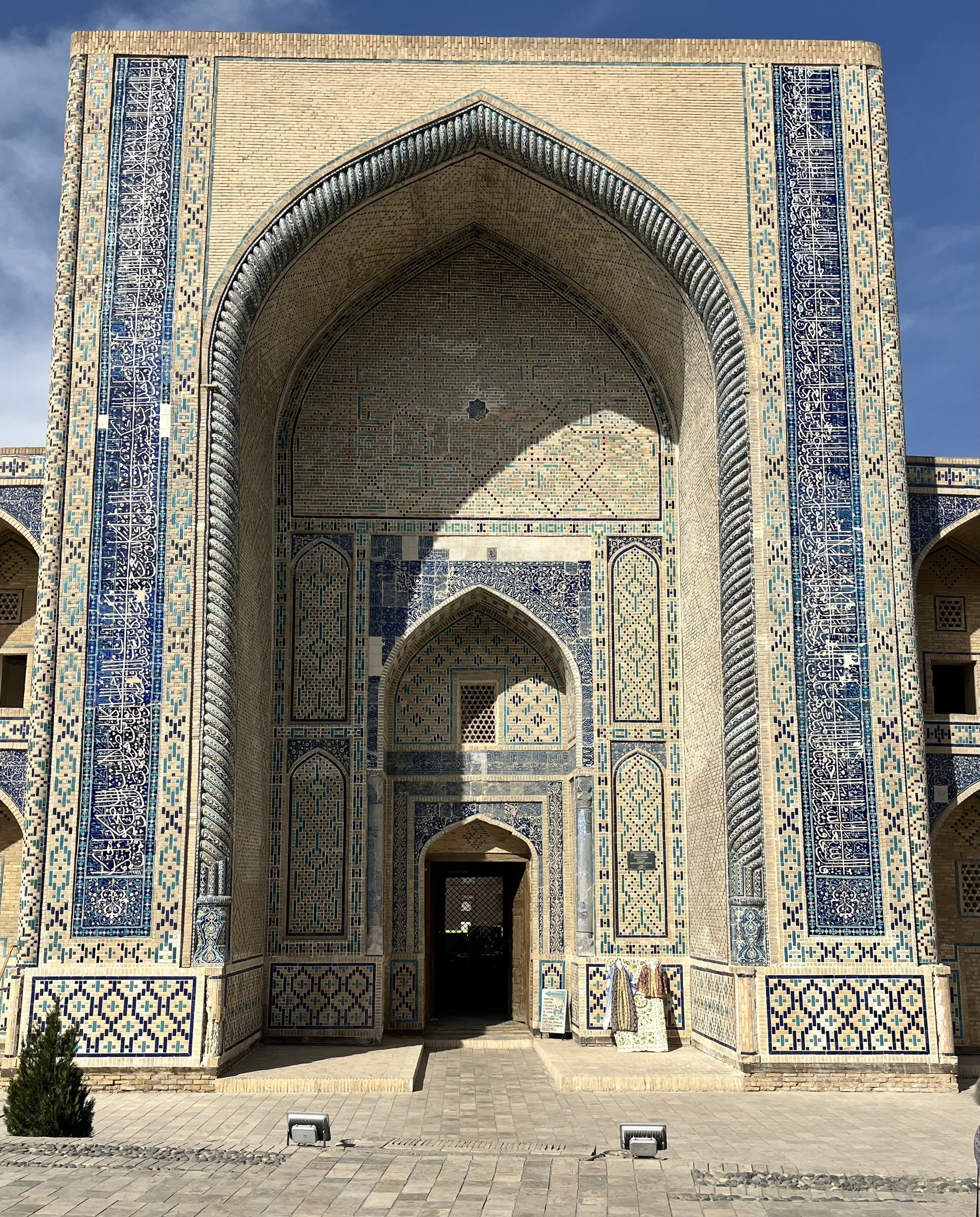 Front view of an Islamic mosque with intricate blue and beige tilework, pointed arches, and a central doorway. The facade features detailed patterns and calligraphy, with sunlight casting shadows on the entrance.