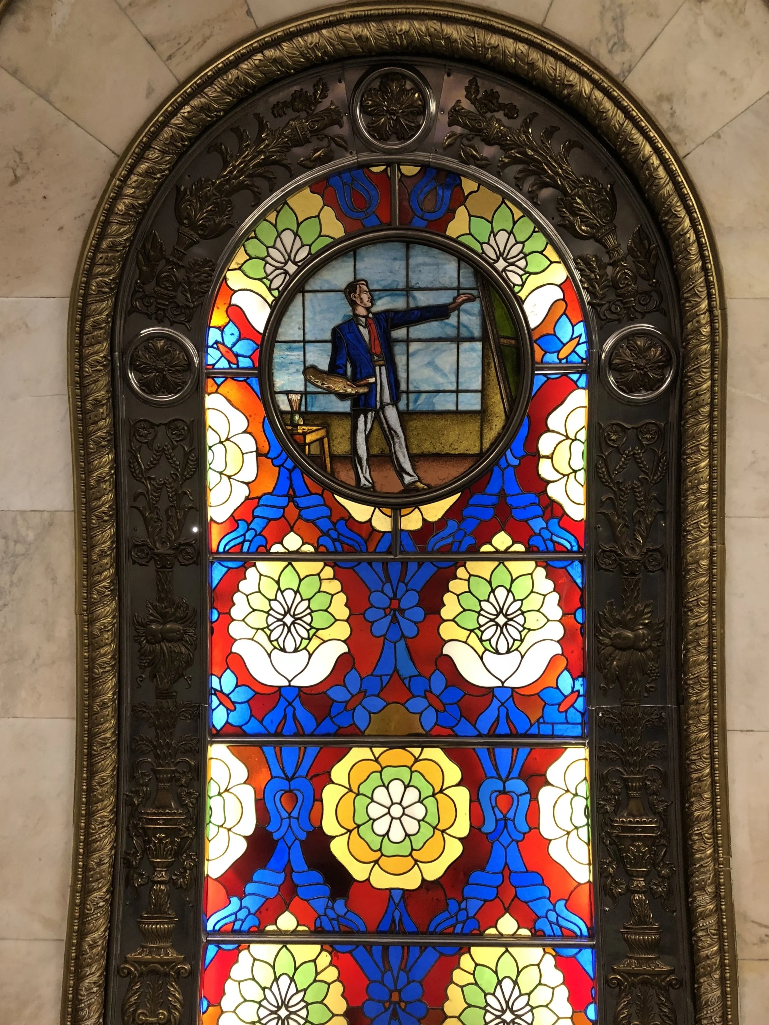 Colorful stained glass window with a man painting at an easel in a room, surrounded by floral patterns.