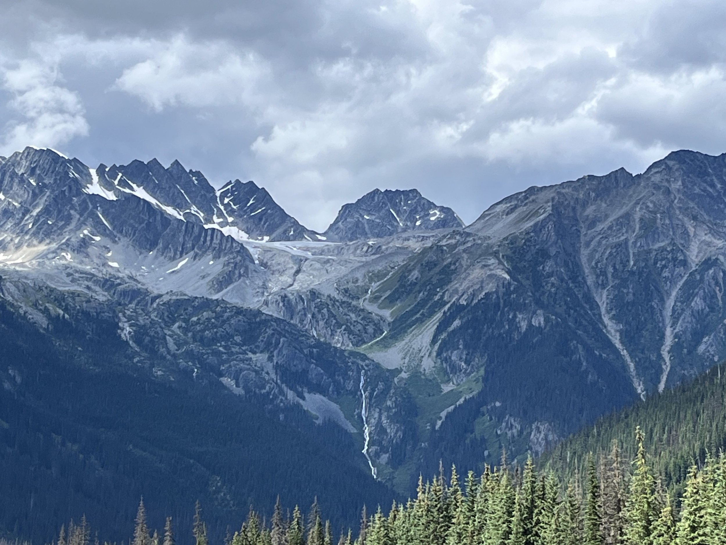 Mountain range with snow patches, green forest in the foreground, and cloudy sky.