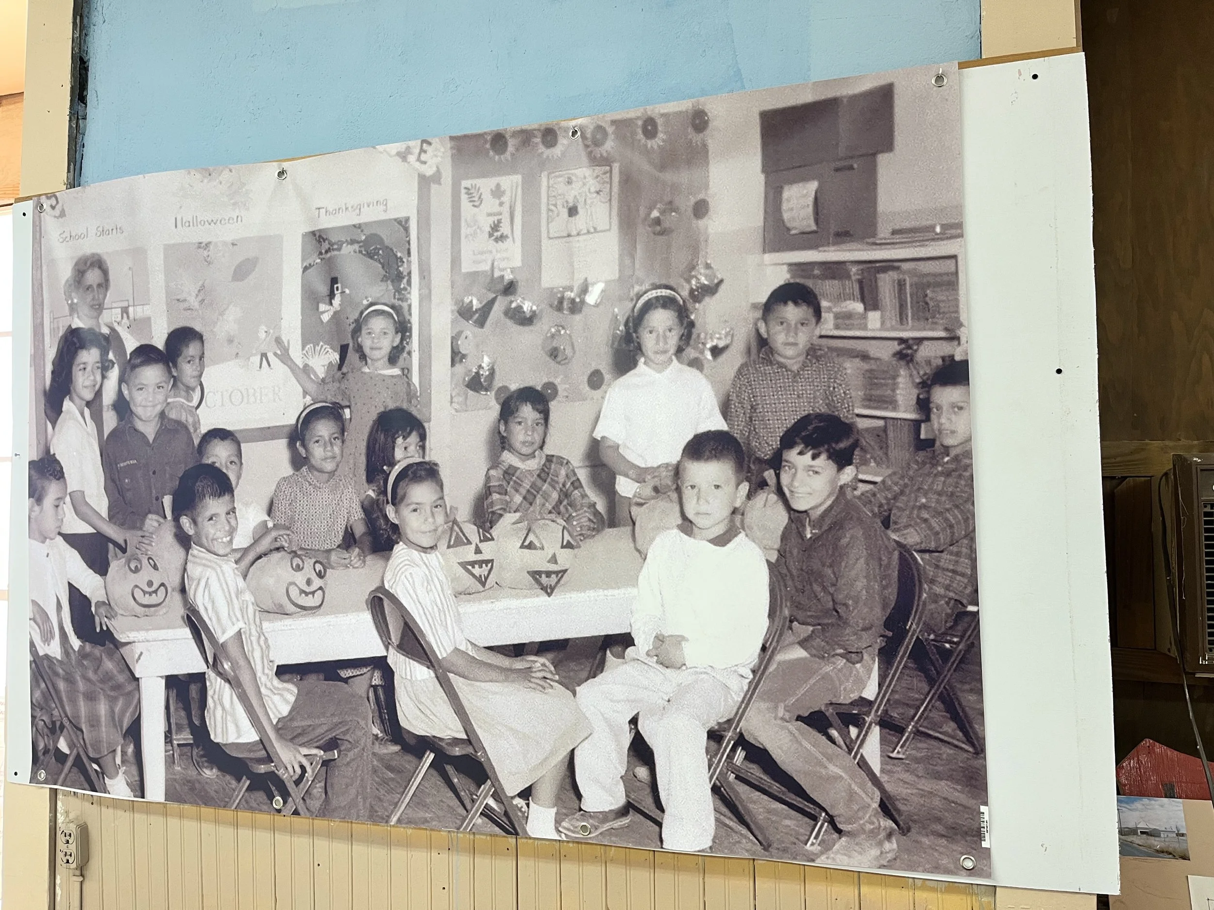 A black-and-white photo of children in a classroom celebrating Halloween, with some children holding pumpkin-shaped bags and there are Jack-o'-lantern decorations on the table, indicating a Halloween party.
