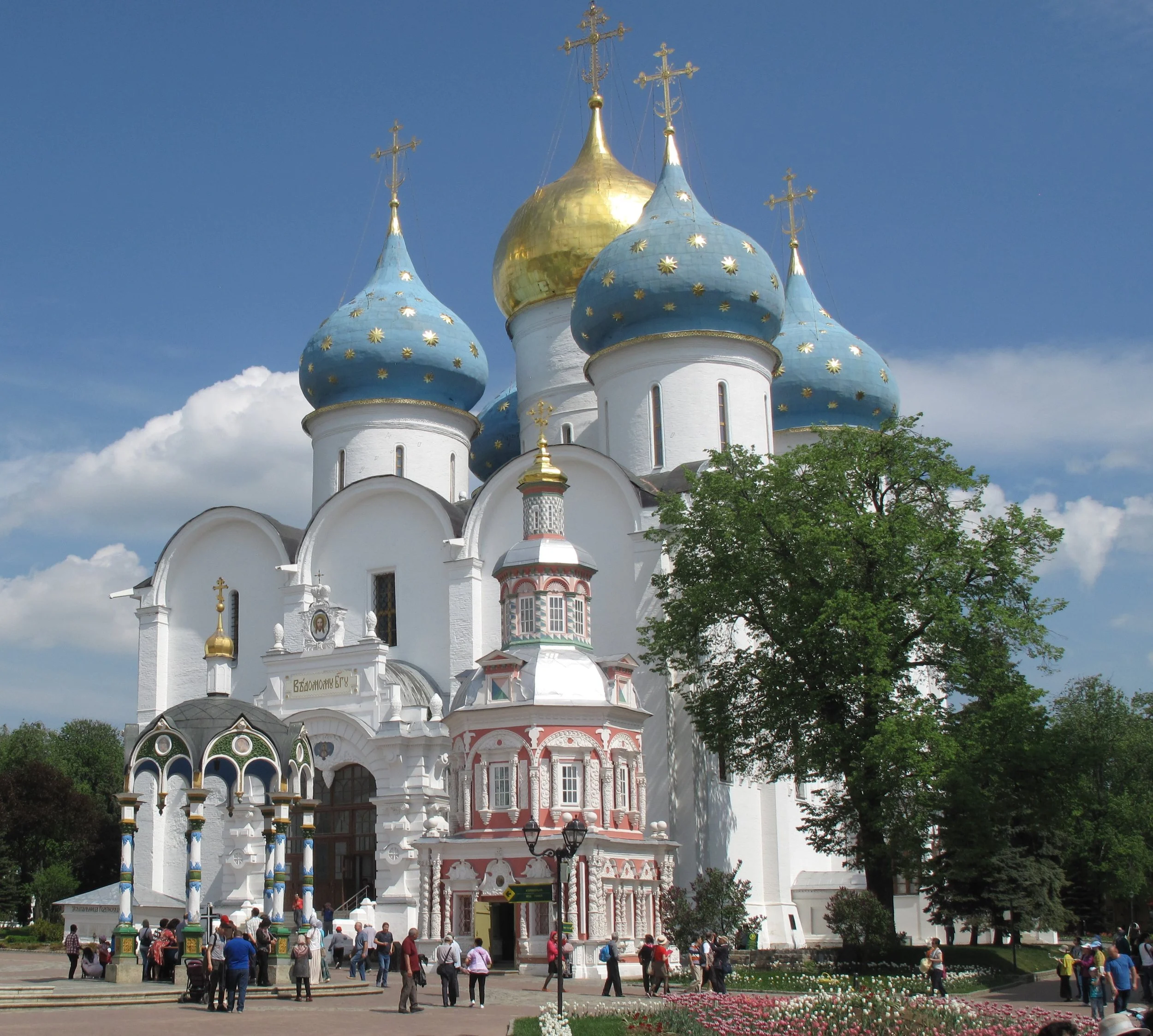 Russian Orthodox church with white walls and blue and gold onion domes, a large green tree in front, and people walking in the courtyard on a sunny day.