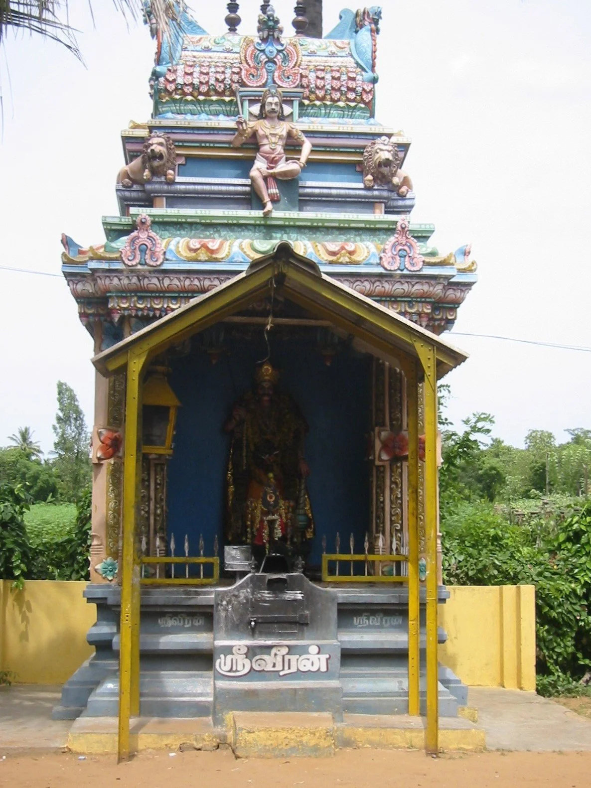 A brightly colored Hindu temple shrine with detailed carvings and statues, featuring a central figure and two lions, set against a backdrop of greenery.