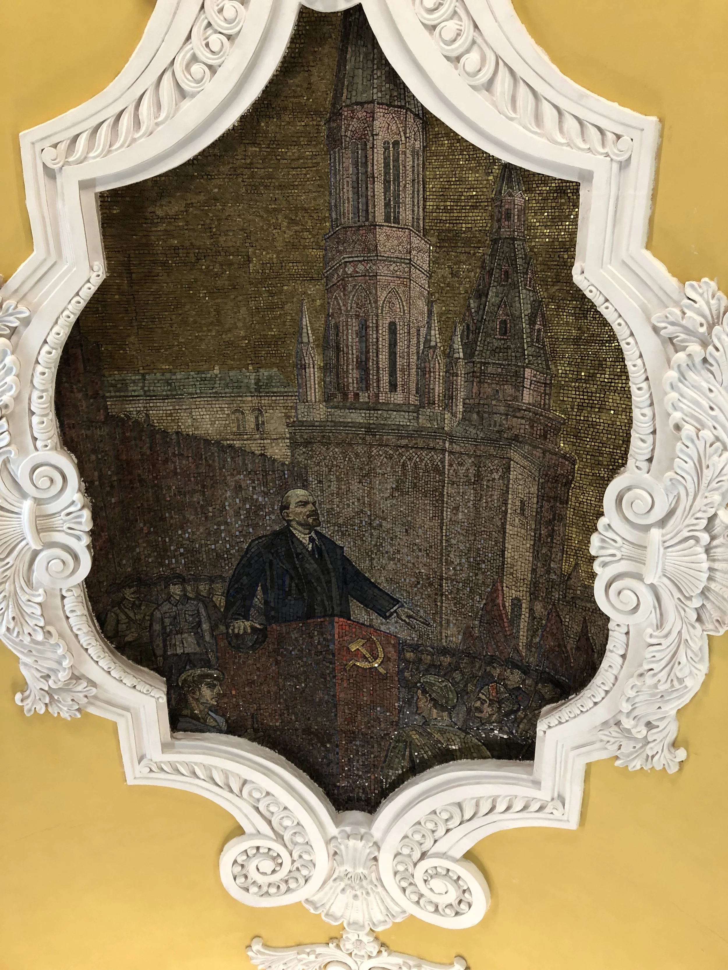 A mosaic of Vladimir Lenin delivering a speech, with a background of a historic building with tall towers, framed by an ornate white decorative border.