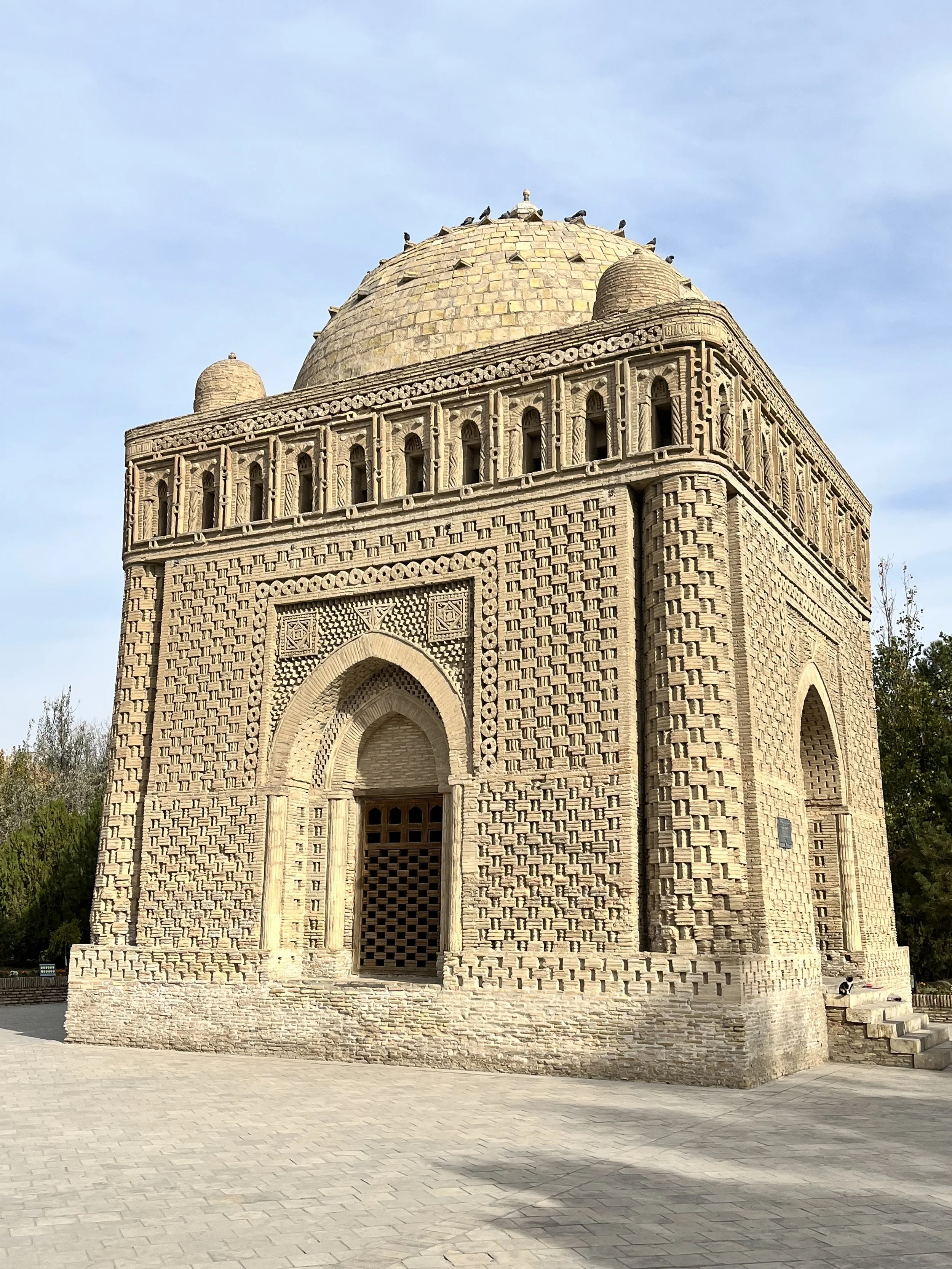 Historic brick mausoleum with a domed roof, arched entrances, and intricate geometric and floral decorations, surrounded by a paved area and greenery.