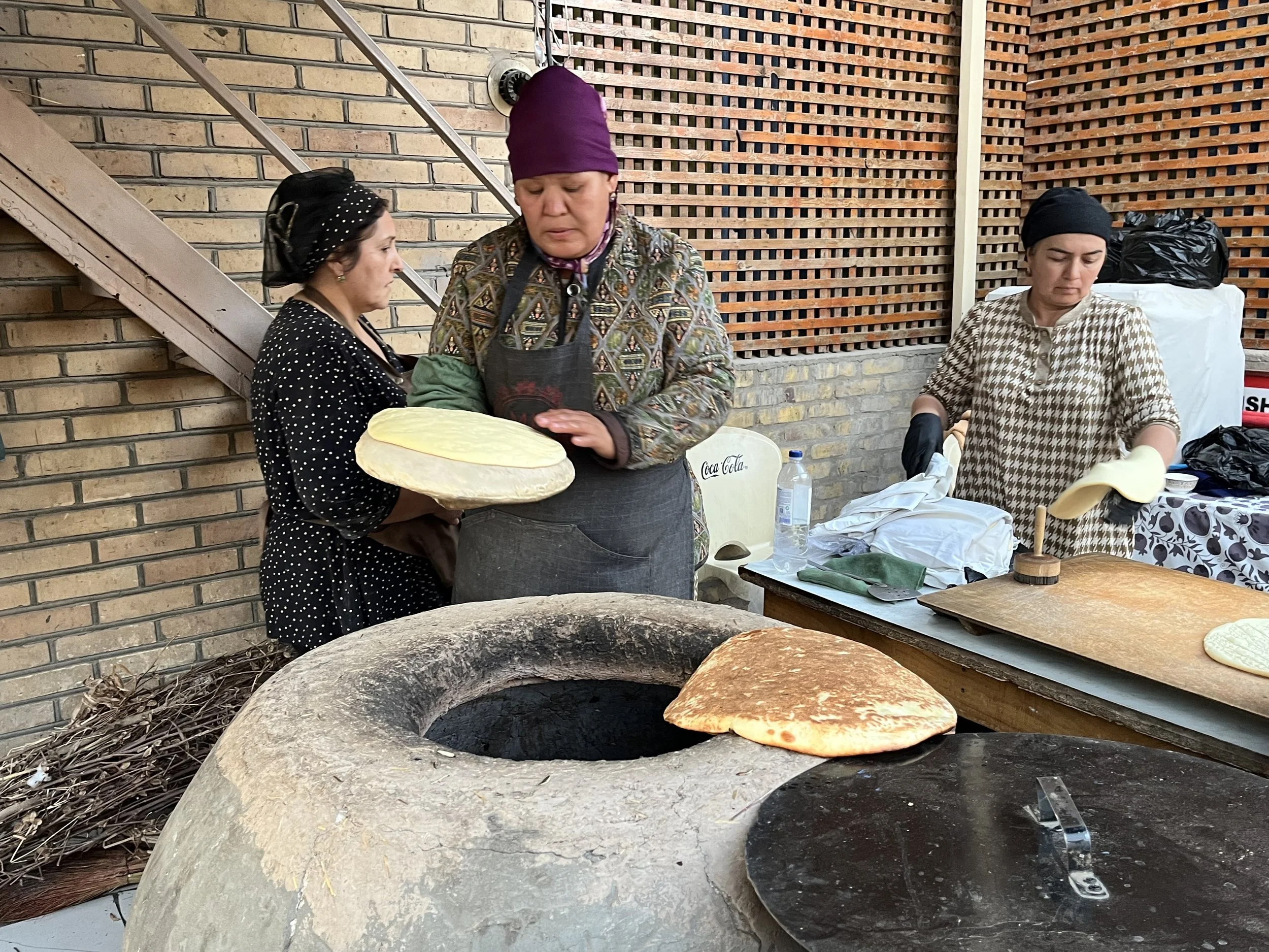 Three women preparing flatbread at an outdoor oven, one woman holding a round piece of dough, the other two working at a table with baked bread and ingredients.